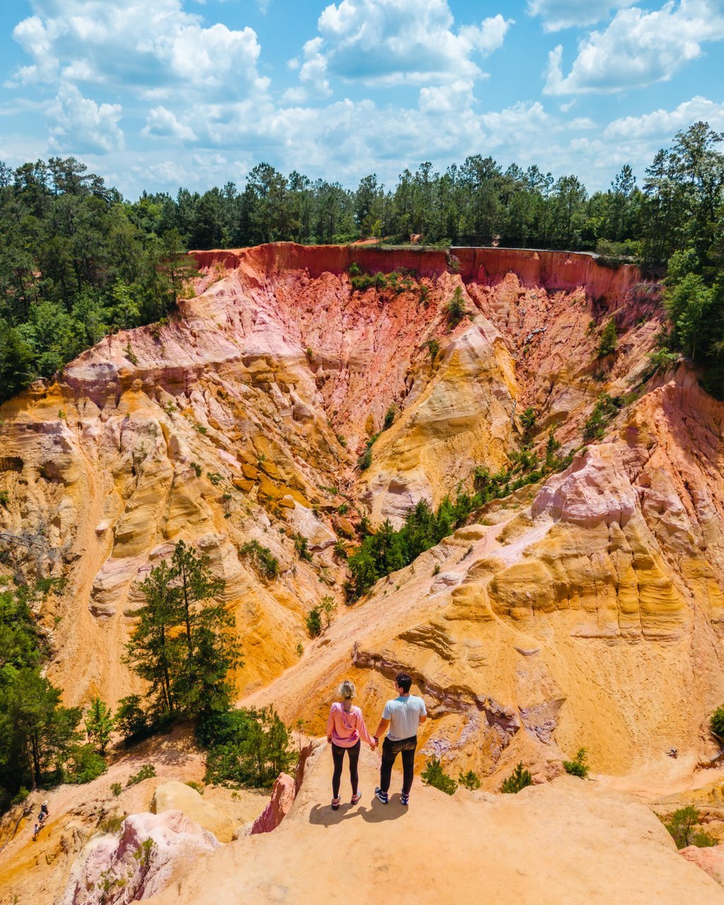 A couple stands on edge of a colorful canyon with yellow, pink, and orange layers, surrounded by green trees and a blue sky with fluffy clouds.