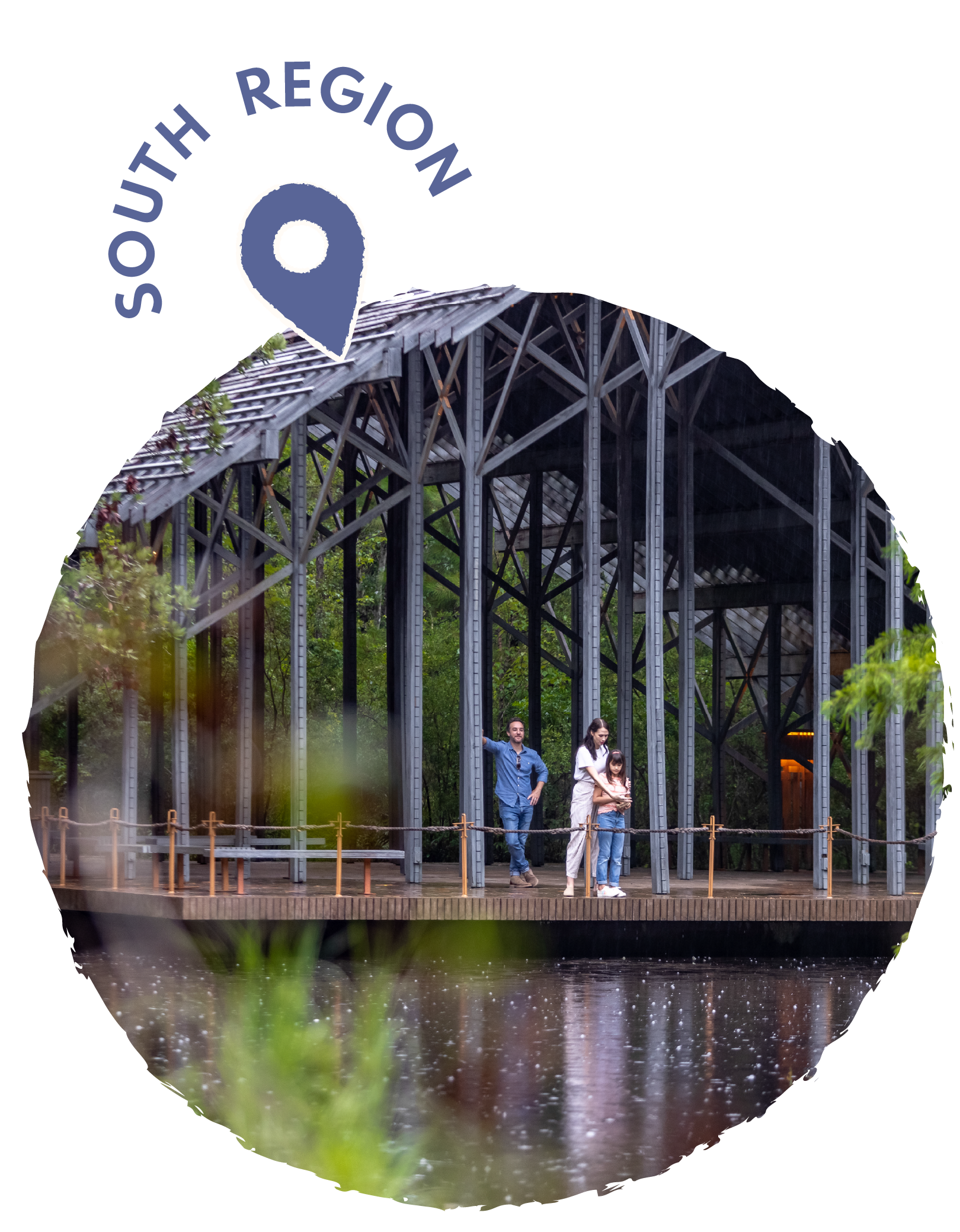 Family of three standing on a wooden platform at a park with a large metal pavilion structure, surrounded by trees and water.