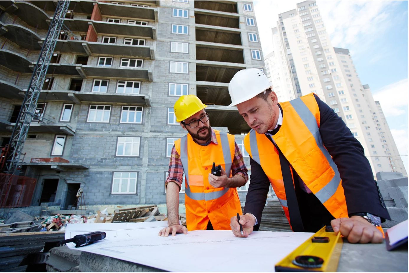 Two construction workers in safety helmets and vests review blueprints on a construction site with an under-construction building and cranes in the background.