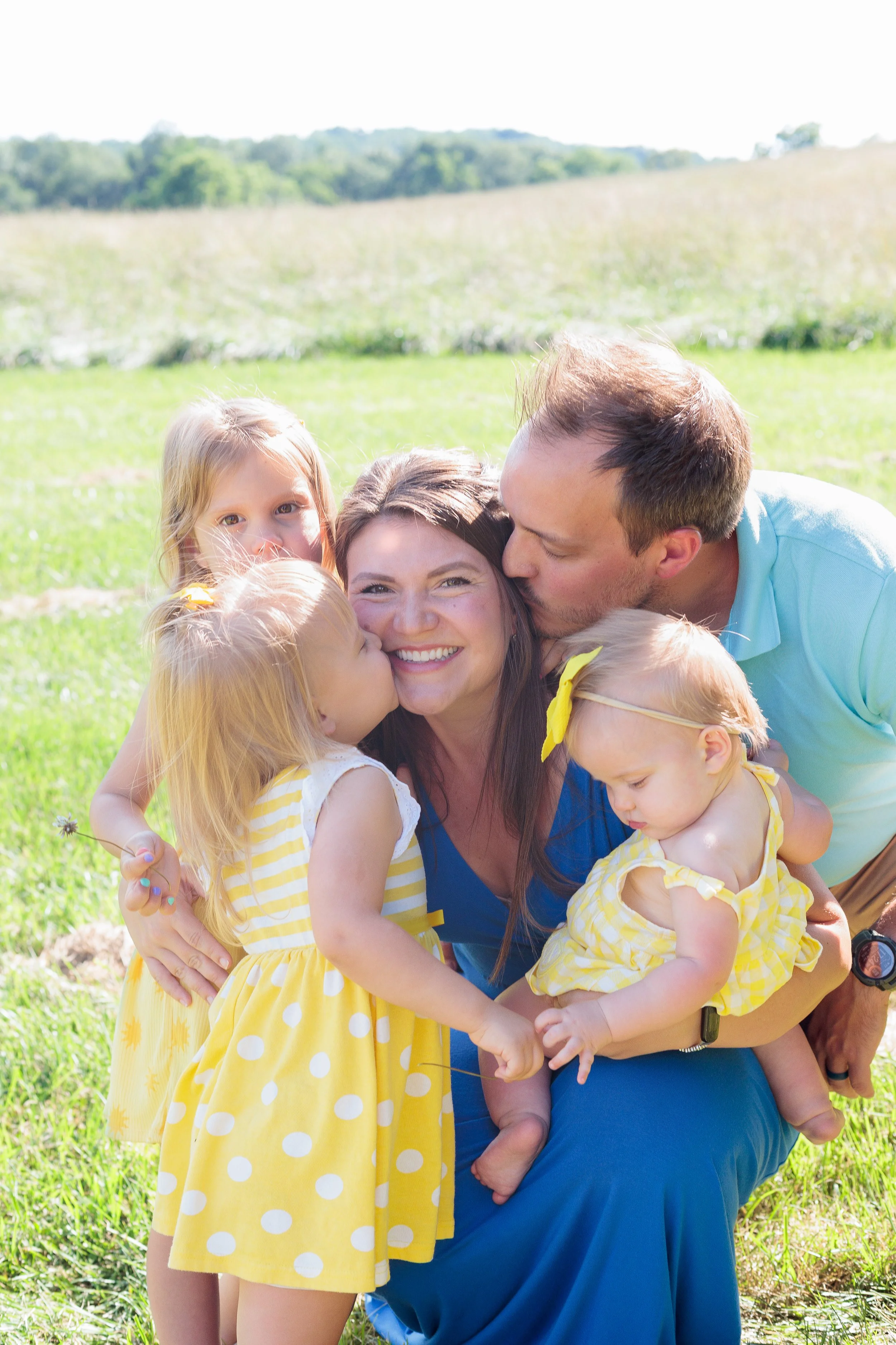 A family outdoors in a grassy field, with a woman, man, and three young children, all wearing yellow and blue clothing, sharing a happy moment.