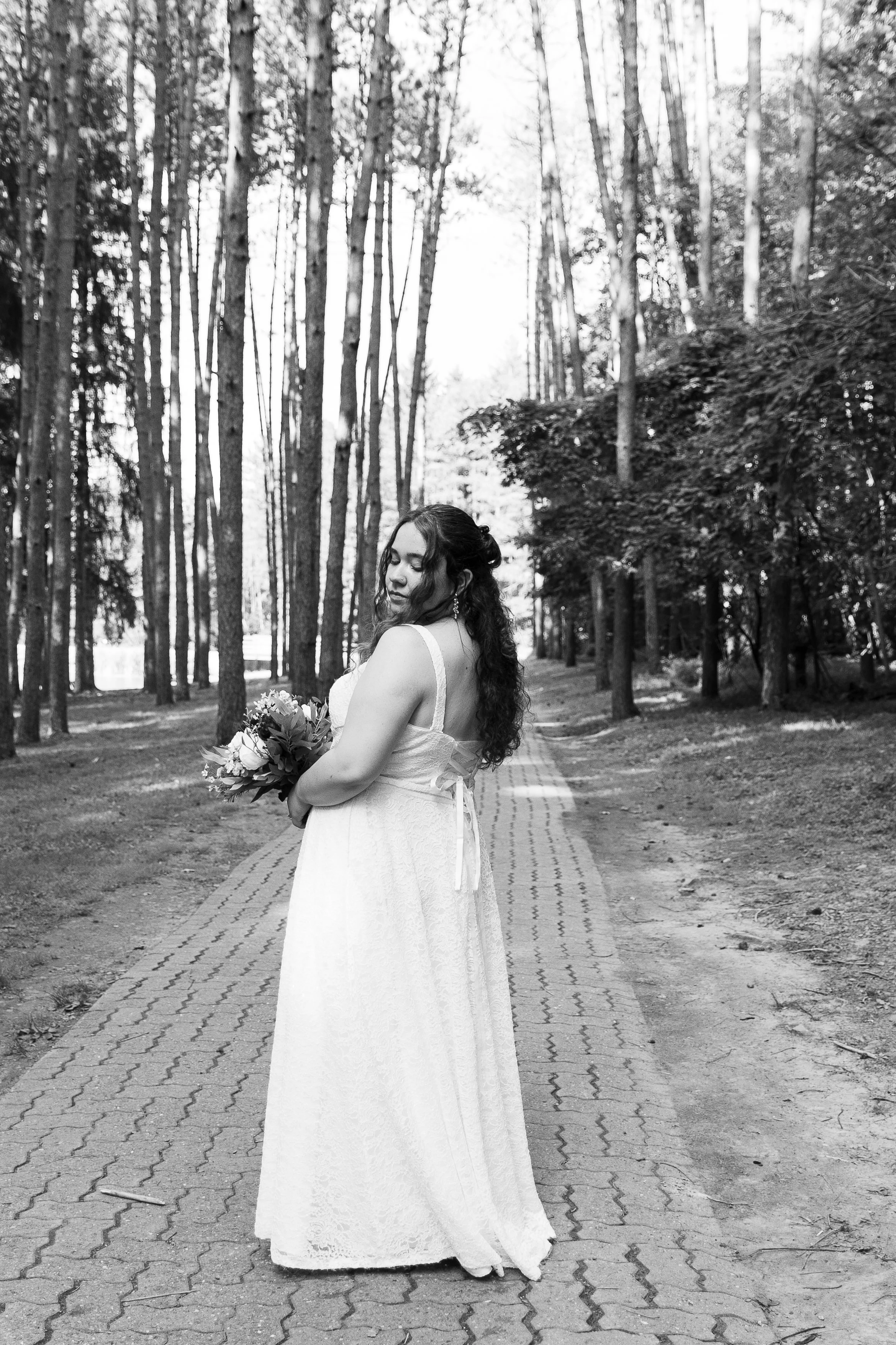A woman in a white dress holding a bouquet standing on a pathway in a forested area, with tall trees surrounding her, captured in black and white.