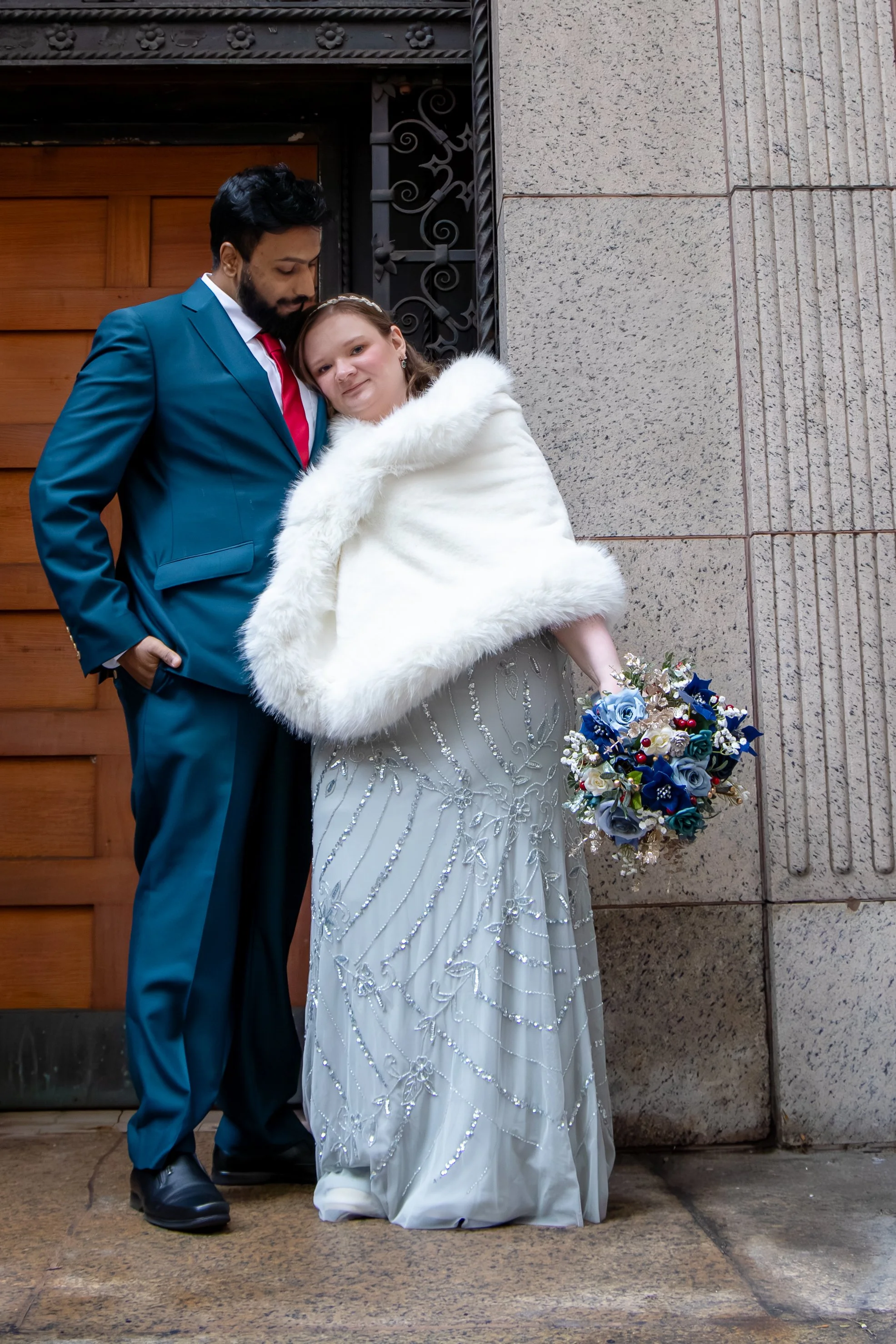 A bride and groom standing close together in a editorial photography outside a building, the bride holding a bouquet of blue flowers and wearing a white gown and coat, while the groom is dressed in a blue suit with a red tie.