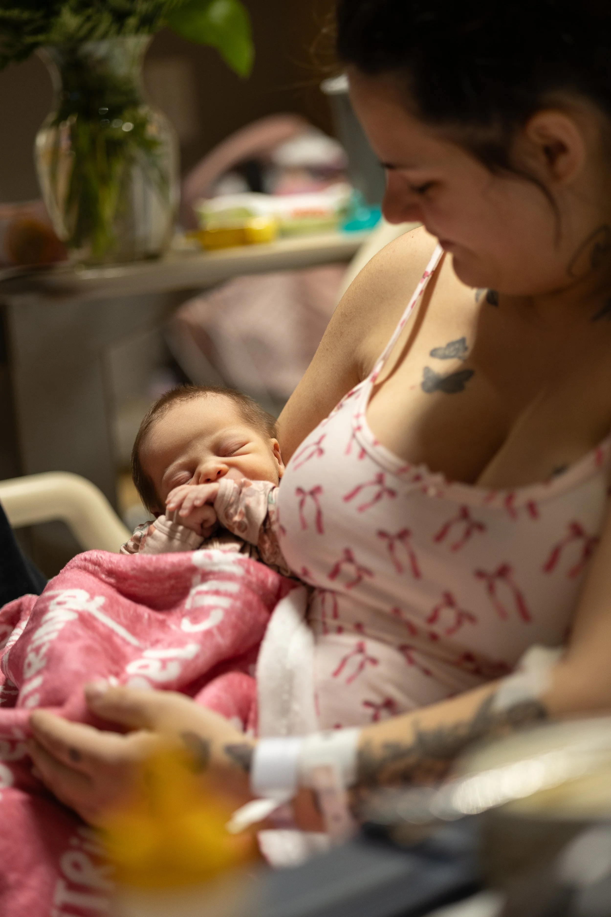 A woman holding a sleeping newborn baby in a hospital room, with medical equipment and flowers in the background.
