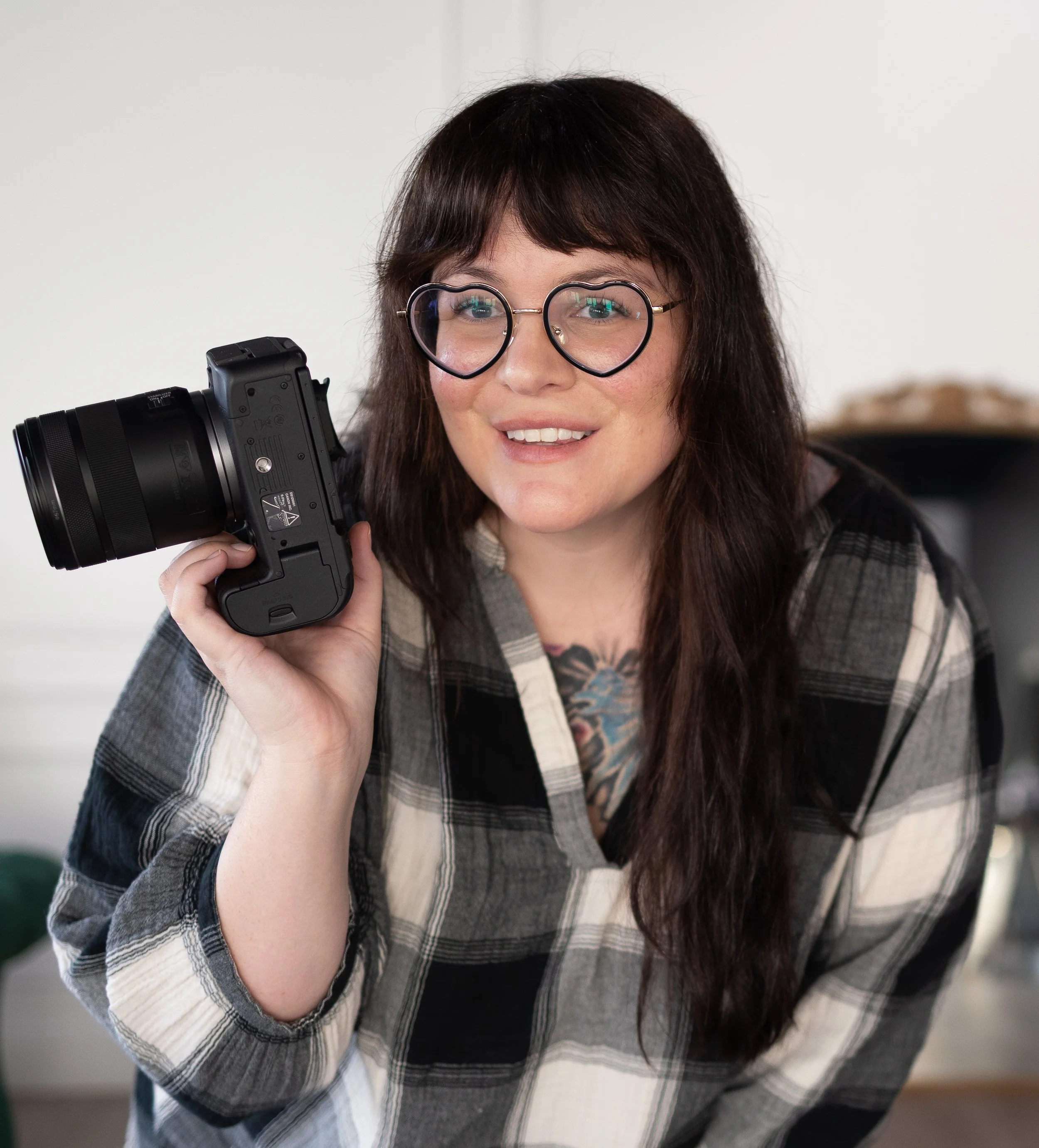 A woman with long brown hair, glasses with heart-shaped frames, and a tattoo on her chest holds a camera and smiles.