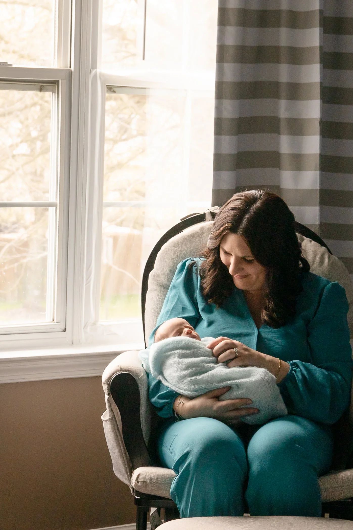 A woman sitting in a chair by the window, holding a newborn wrapped in a blanket, looking at the baby with affection.