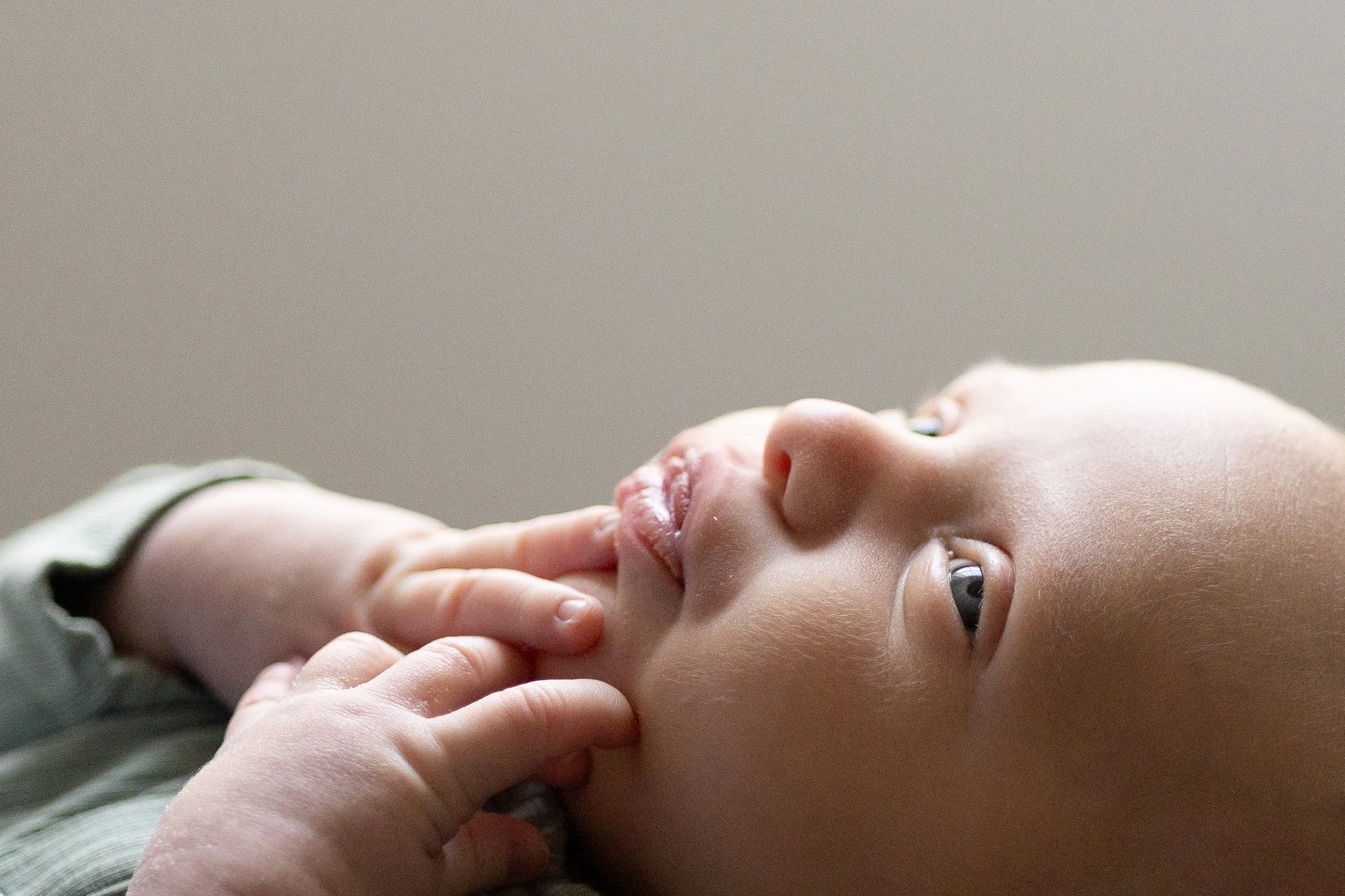 Close-up of a baby lying on its back with one hand touching its chin, looking up with wide eyes, soft lighting, neutral background.