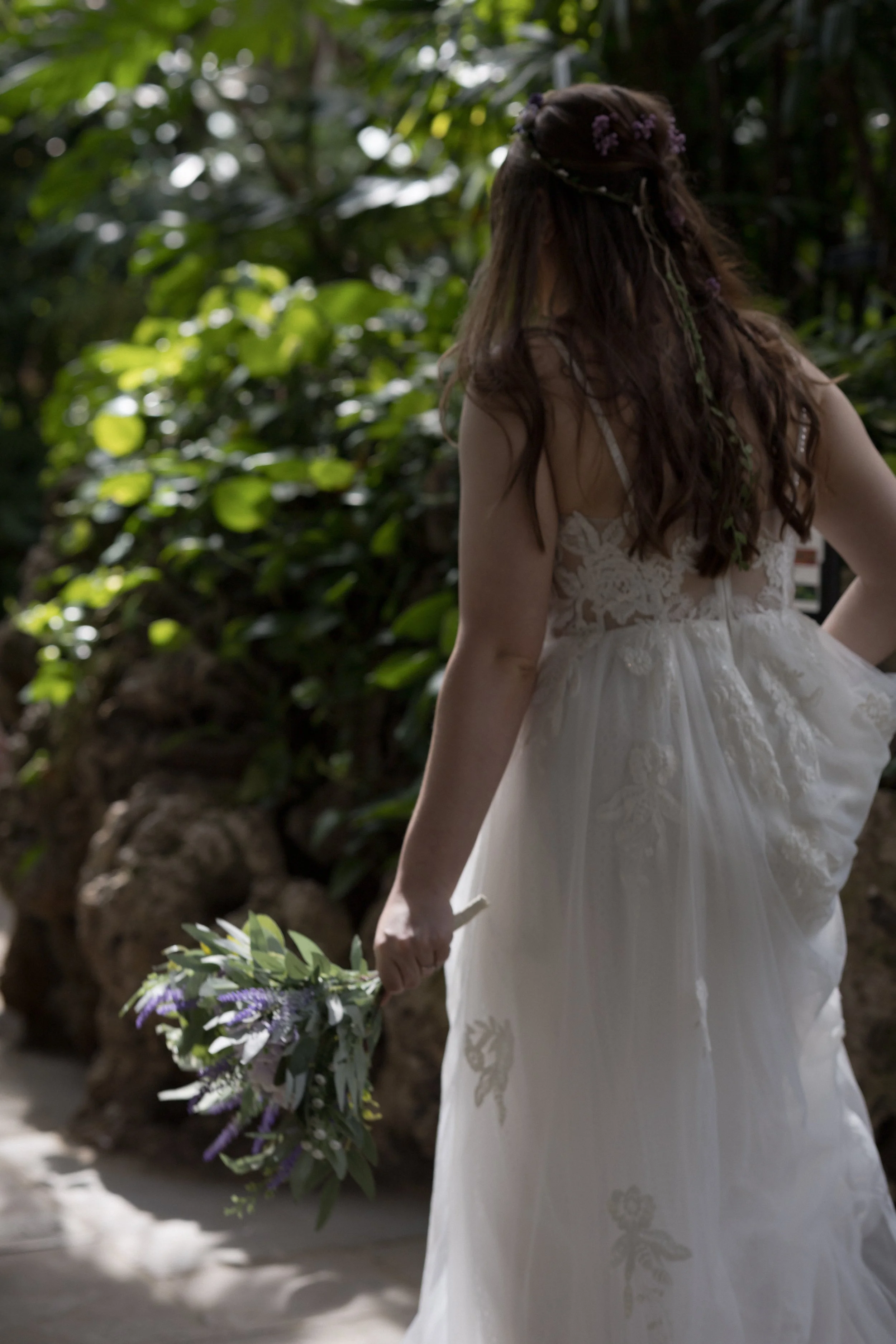 A woman in a white wedding dress holding a floral bouquet, walking outdoors near lush green foliage.