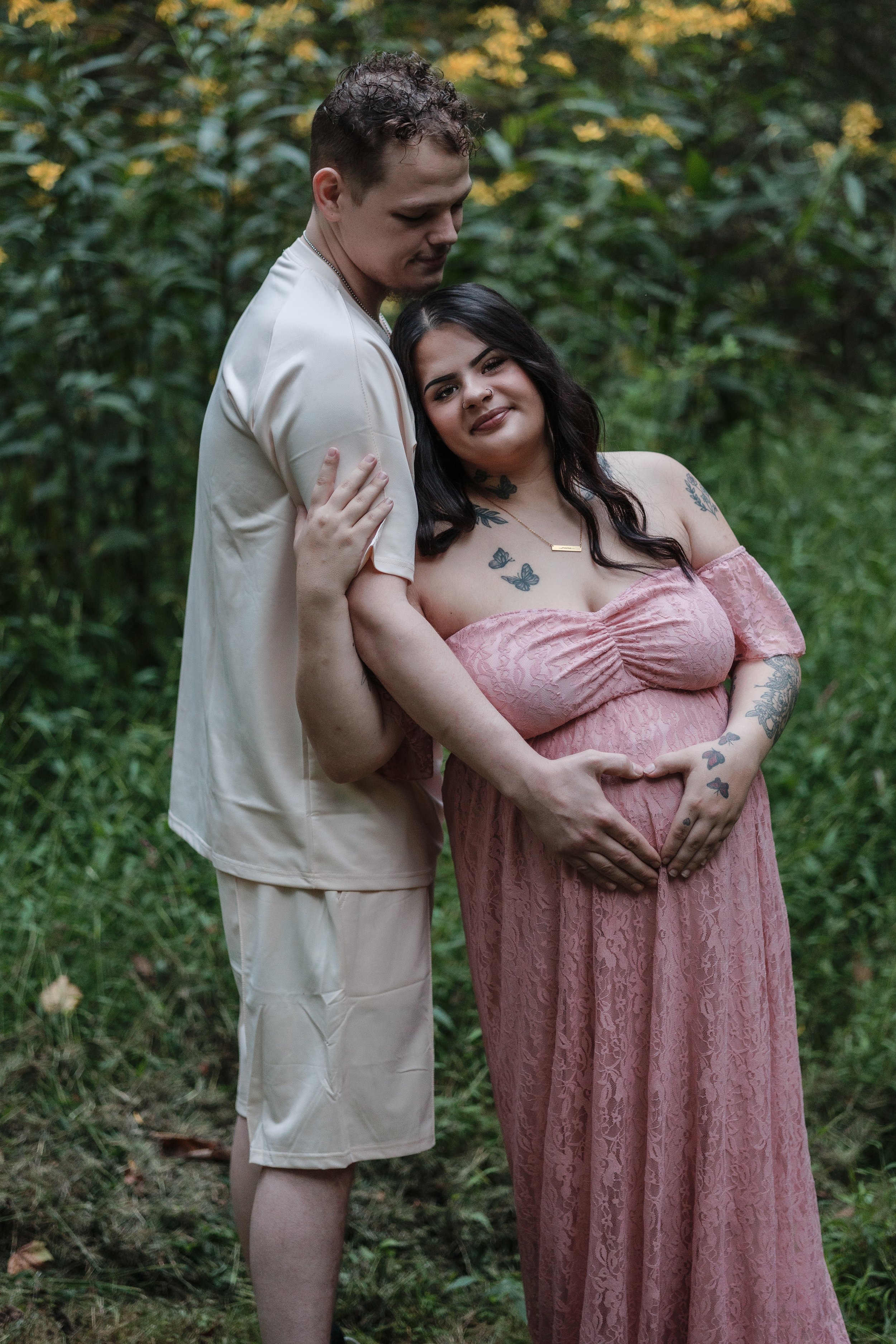 A pregnant woman with dark hair and tattoos on her arms and chest in a pink off-shoulder dress, standing next to a man in beige clothing, both embracing in a green outdoor setting ethereal maternity photography session in Canonsburg PA 