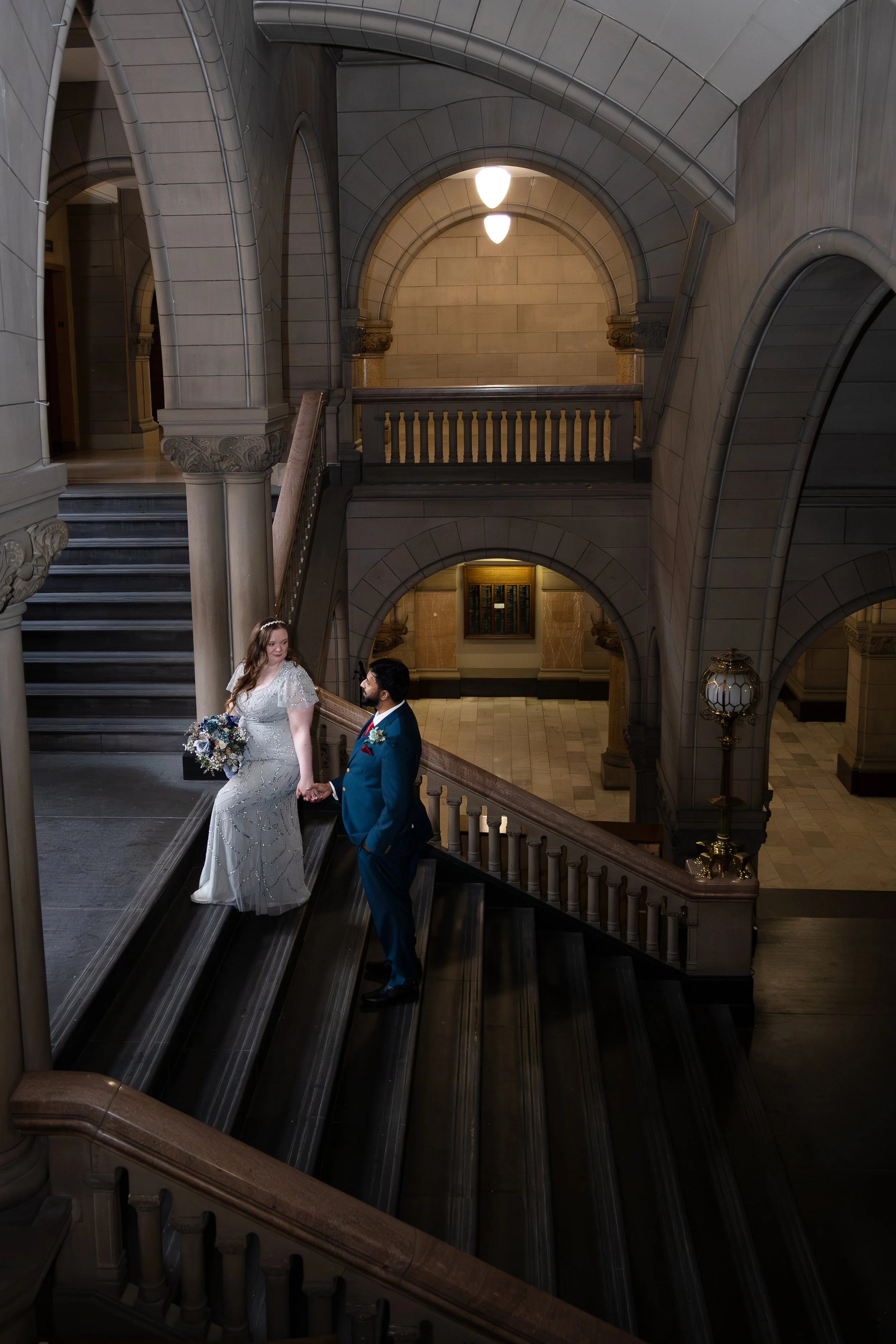 A bride and groom photography stand on the stairs of an ornate, historic building with high arch ceilings, holding hands and gazing at each other in a moody documentary style photograph at the Allegheny County Courthouse Elopement in Pittsburgh