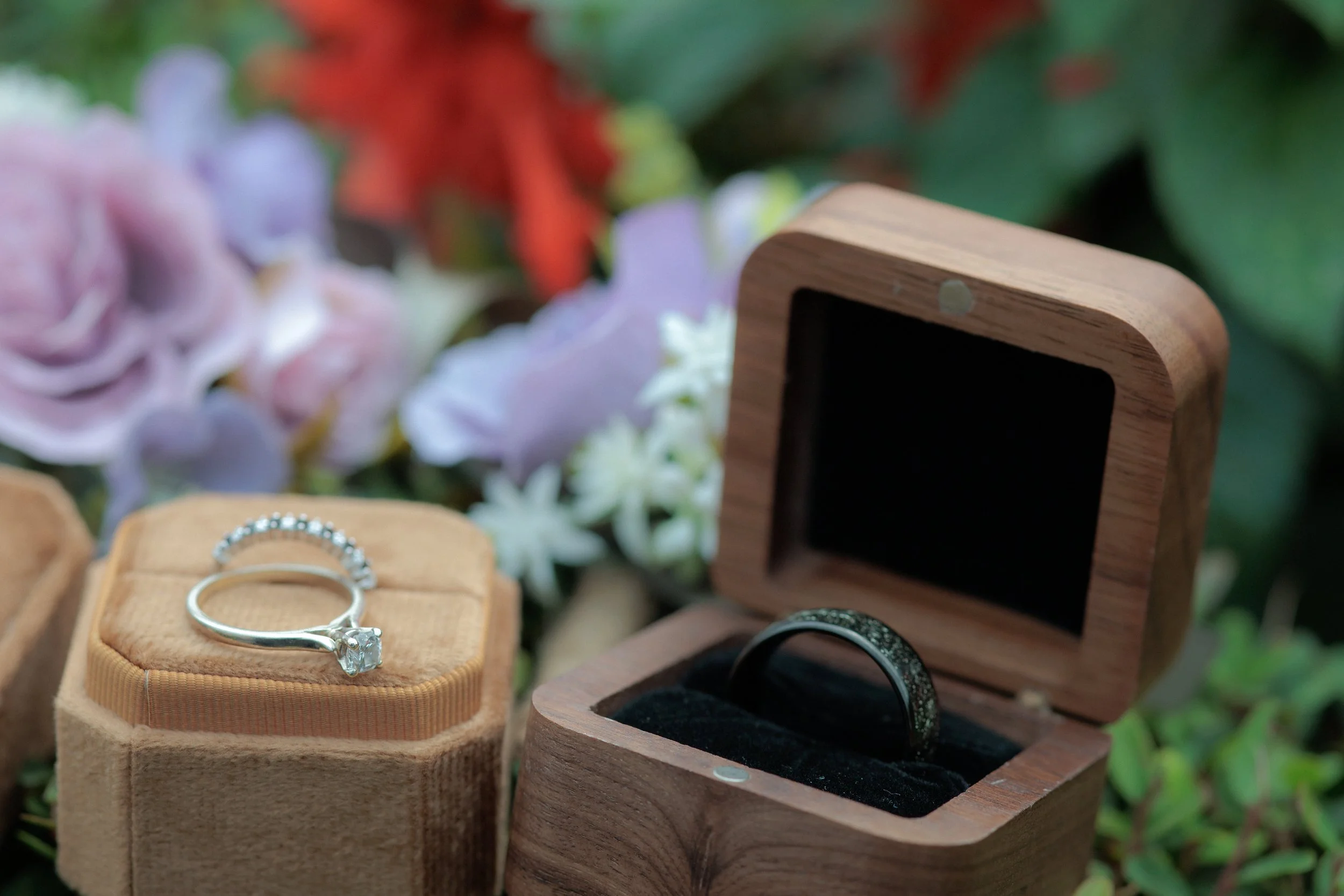Engagement ring with a large center diamond on a beige velvet ring box, and a black wedding band with small diamonds in a wooden ring box, surrounded by colorful flowers.