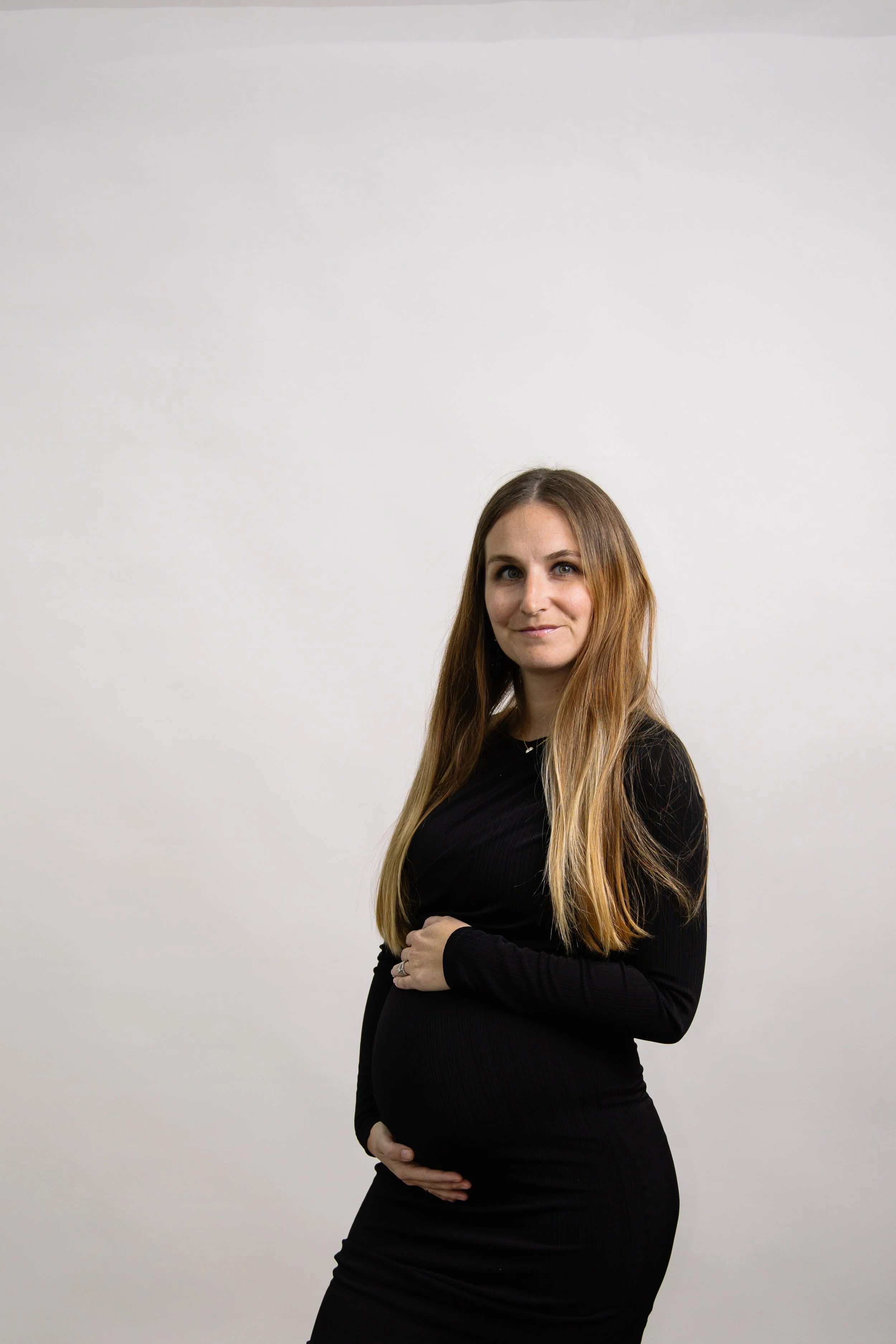 A pregnant woman with long, wavy, light brown hair, wearing a black long-sleeve dress, stands facing partly to the right, cradling her belly with her right hand and looking at the camera against a plain white background.