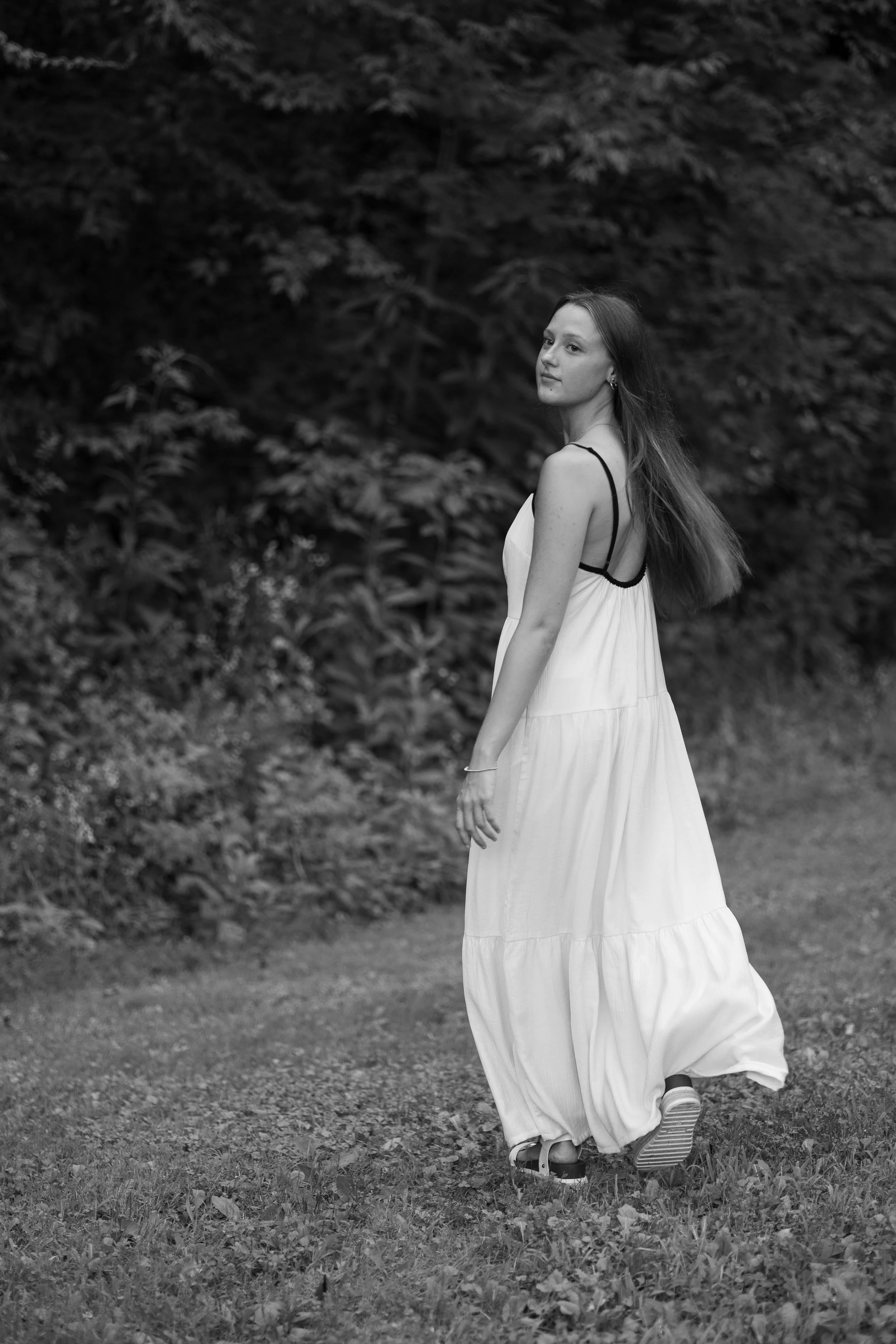 A young woman in a long white dress walking outdoors on a grassy path with dense foliage in the background.