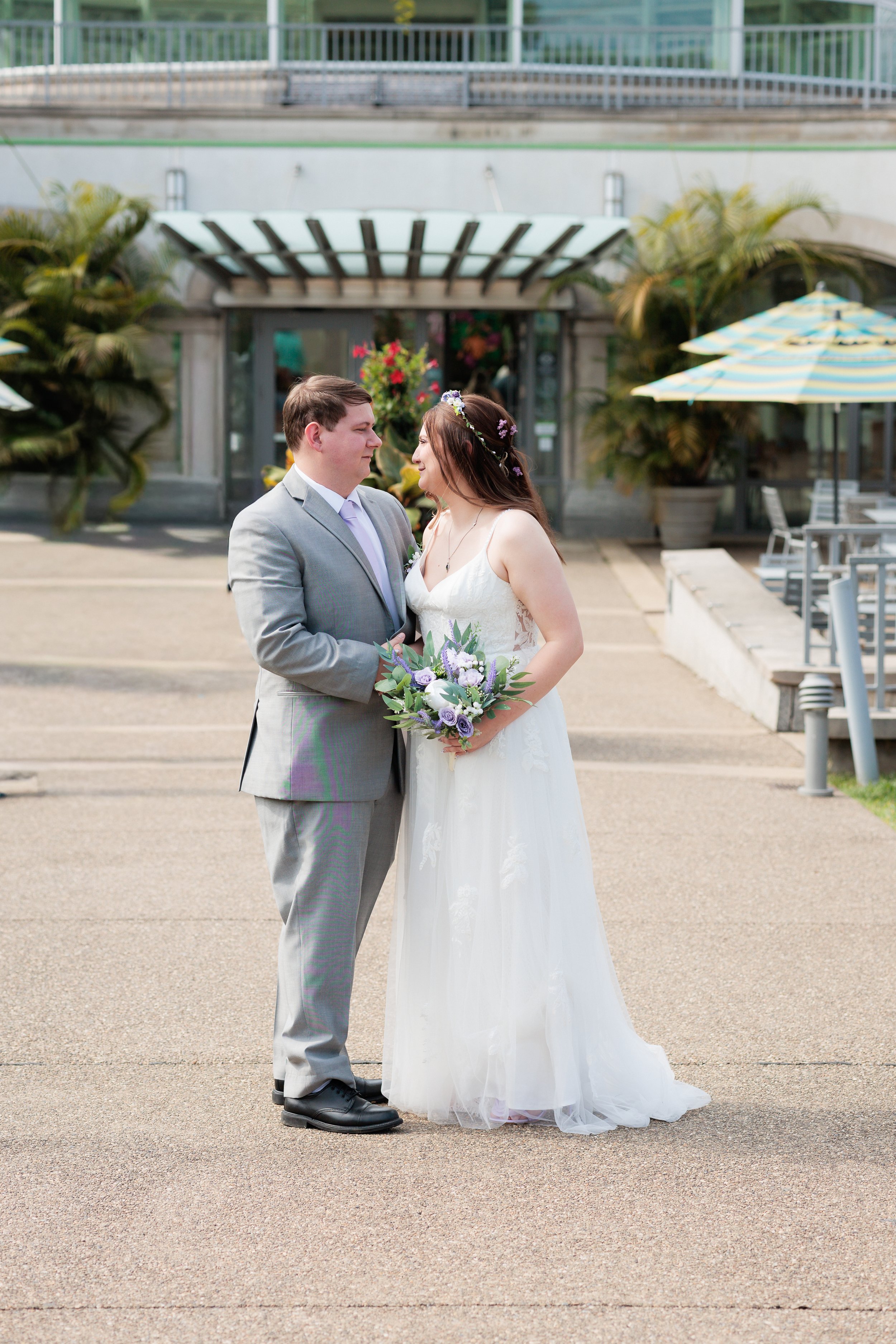 A bride and groom standing close together, holding hands, outside a building with tropical plants, umbrellas, and outdoor seating, on their wedding day.
