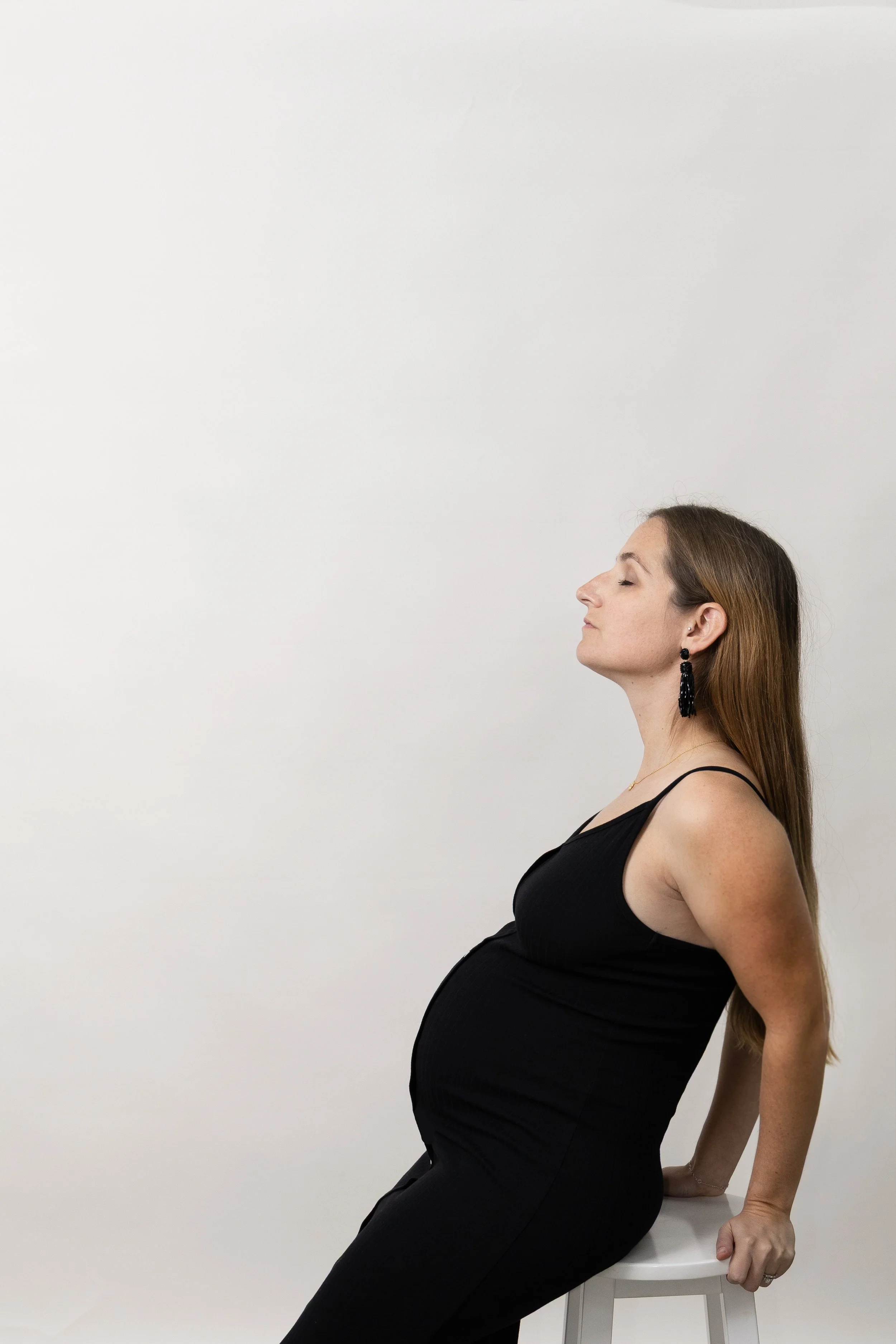 A pregnant woman with long brown hair wearing a black sleeveless dress and black earrings, sitting on a white stool with her eyes closed against a plain white background.