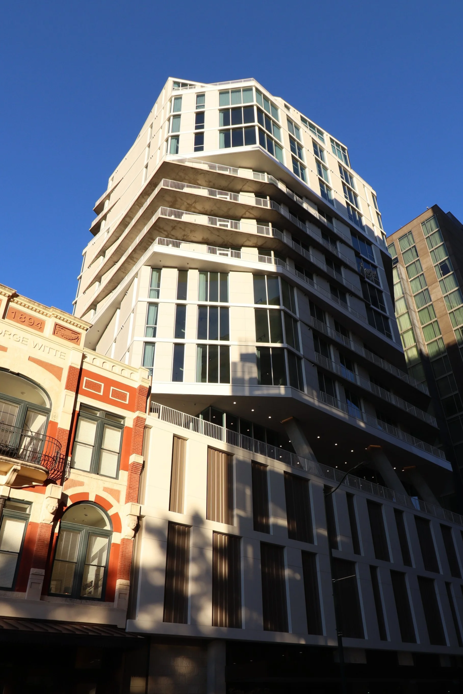 Modern multi-story building with glass windows and balconies, located next to an older building with decorative brickwork, under a clear blue sky.
