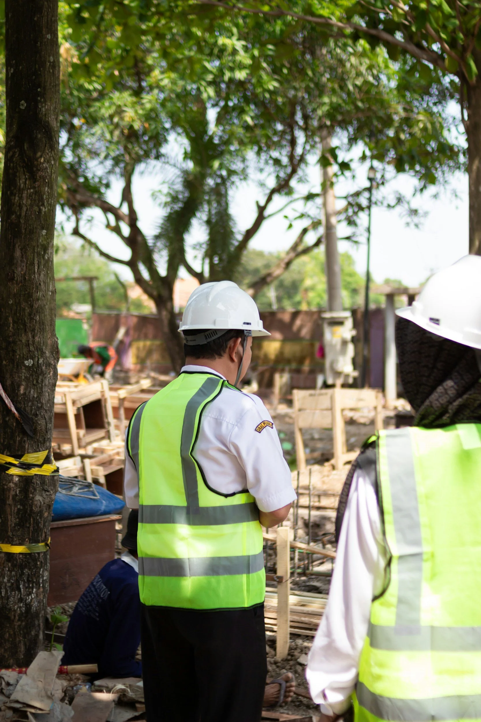 Construction workers wearing safety helmets and reflective vests on a construction site with trees and wooden structures in the background.