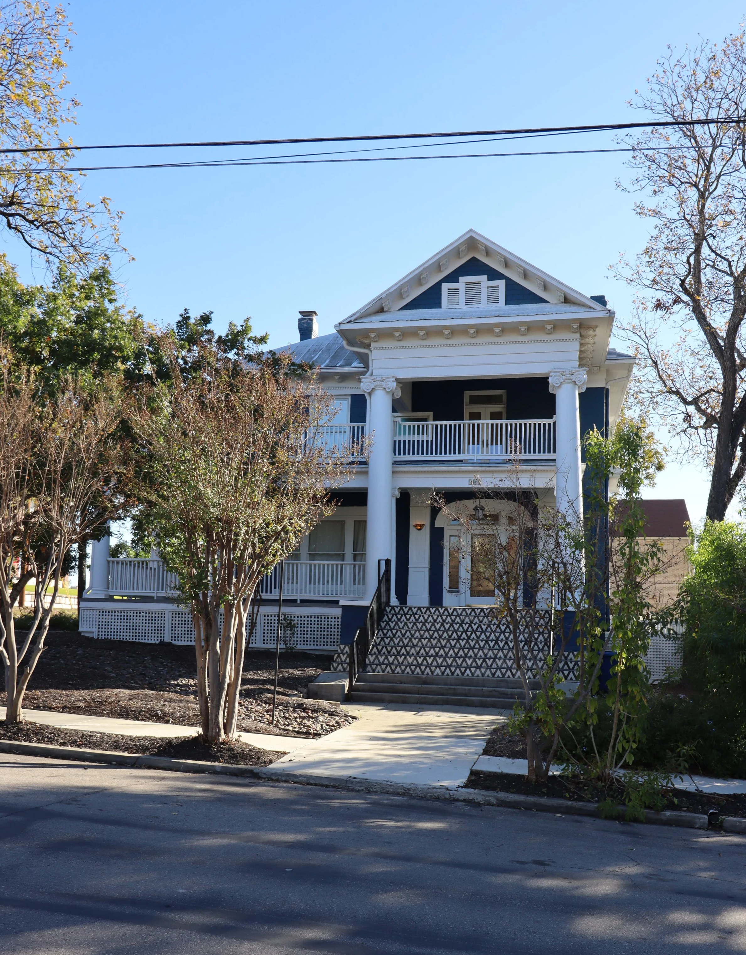 A two-story house painted blue with white trim, featuring a front porch, columns, a staircase, and a balcony, surrounded by trees and a paved sidewalk.