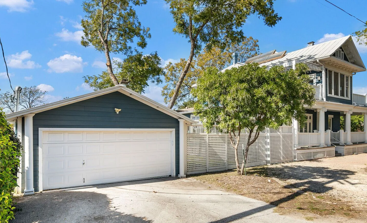 A suburban house with a white porch and multiple windows, a detached garage, trees, a partly cloudy sky, and a driveway.