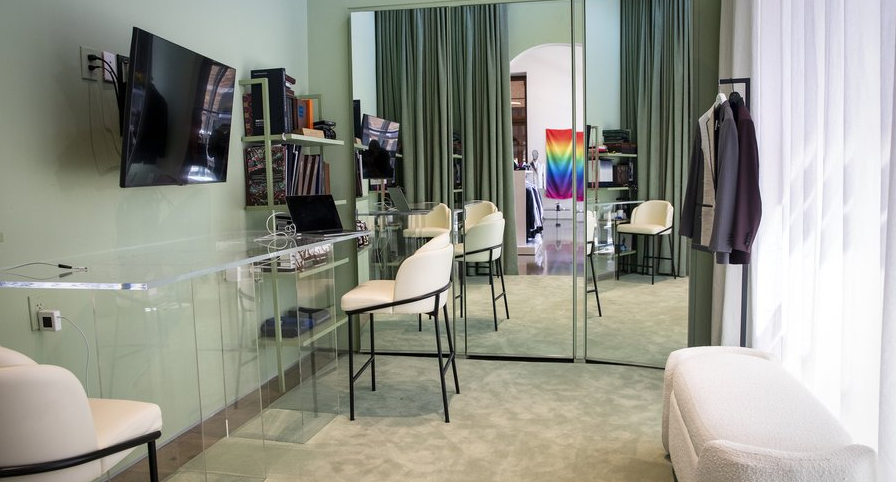 A modern room with a glass desk, white chairs, a wall-mounted TV, and a mirrored sliding door leading to another room with a rainbow flag and a clothing rack. Green curtains cover large windows, and the room has light-colored walls and carpet.