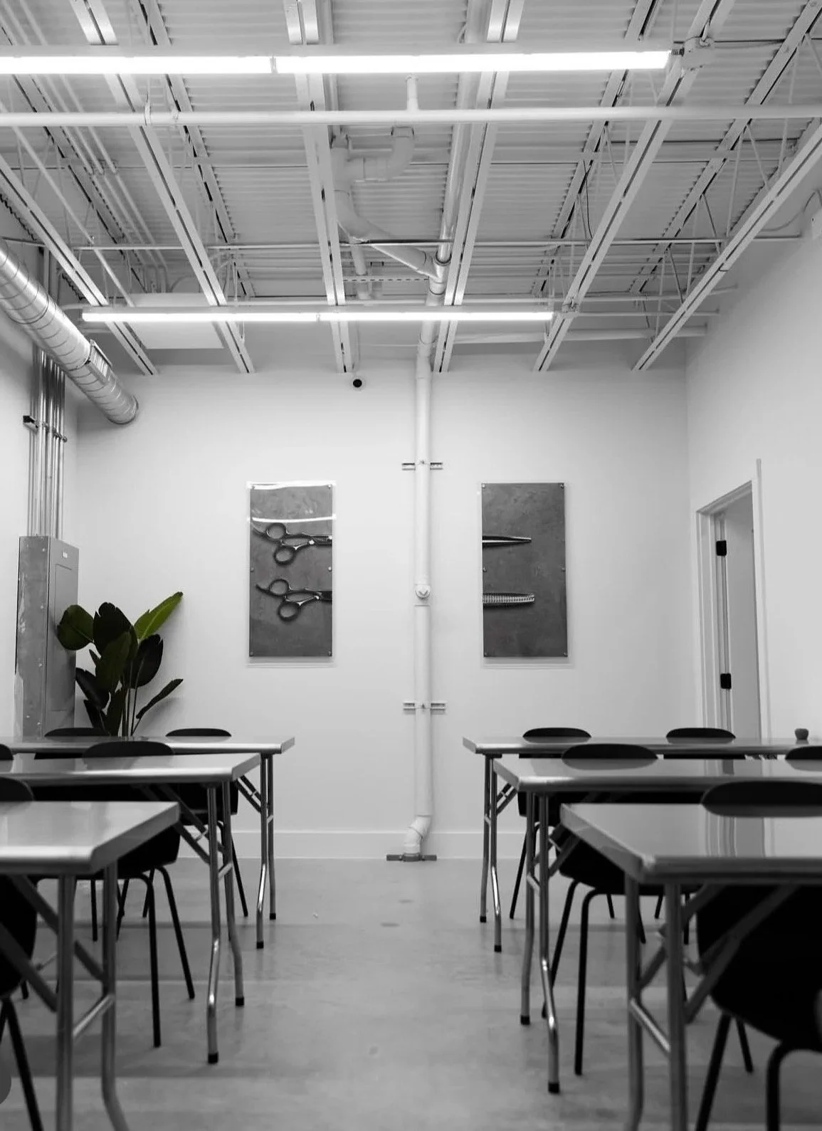 Empty classroom or meeting room with tables and chairs. Two frames with scissors and a pencil on the wall. A plant in the corner, and an exposed ceiling with pipes and lighting.