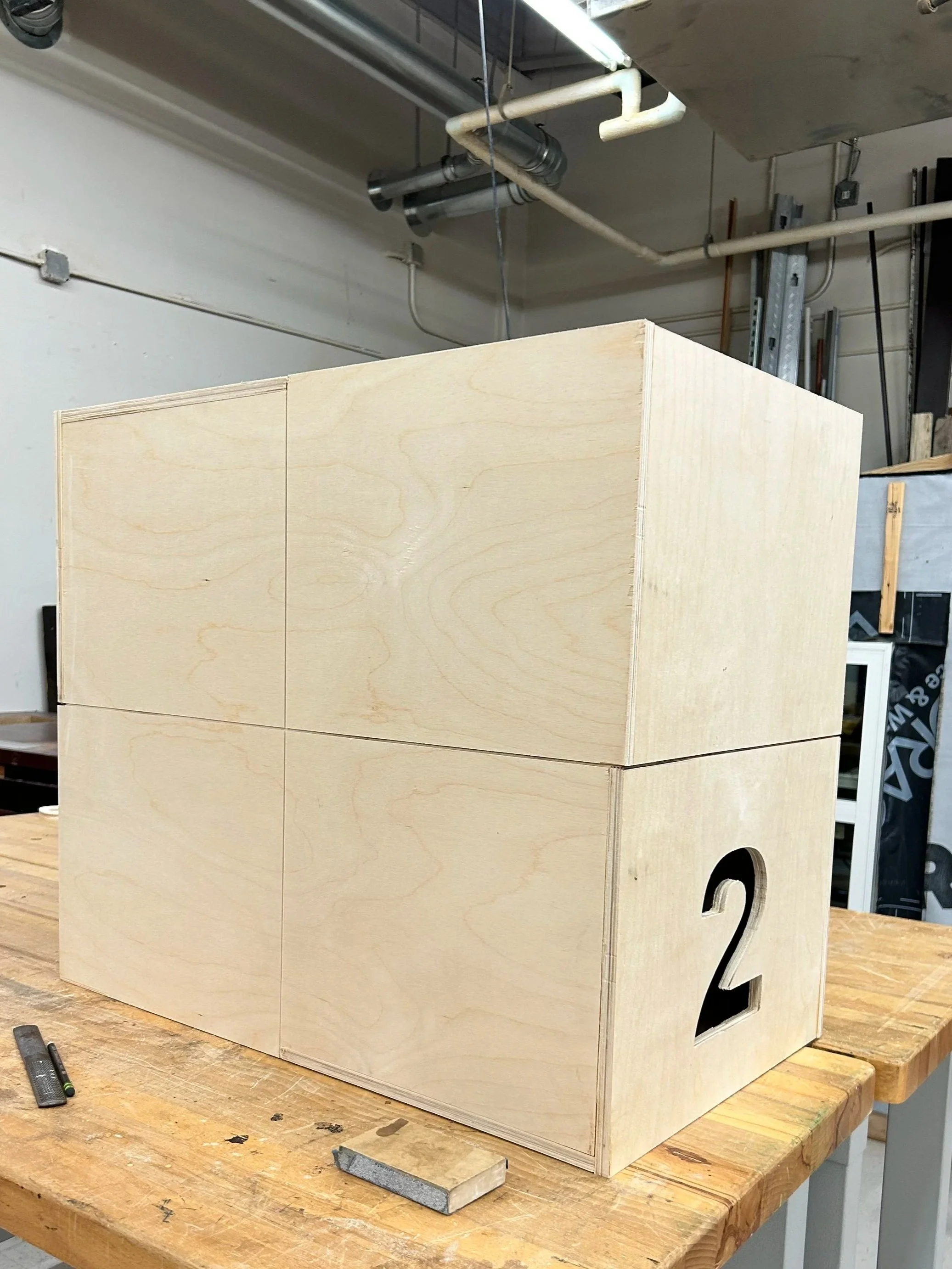 A wooden cube with the number 2 cut out on one side, placed on a wooden workbench in a workshop.