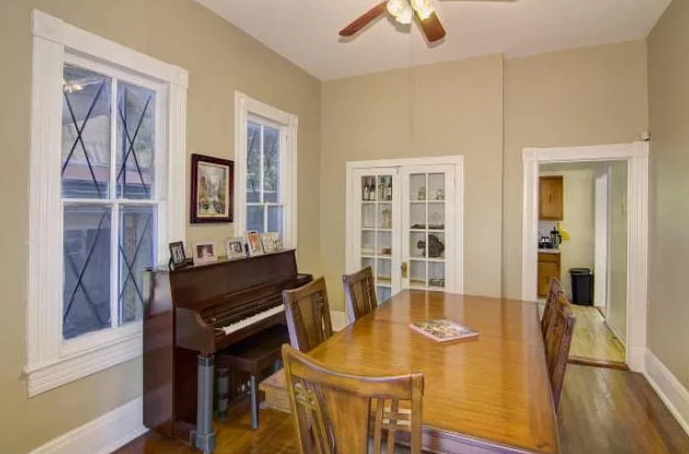 A dining room with a wooden table and six chairs, a piano against the wall with framed pictures on top, two windows, and a glass-paneled door leading to another room with a kitchen visible.