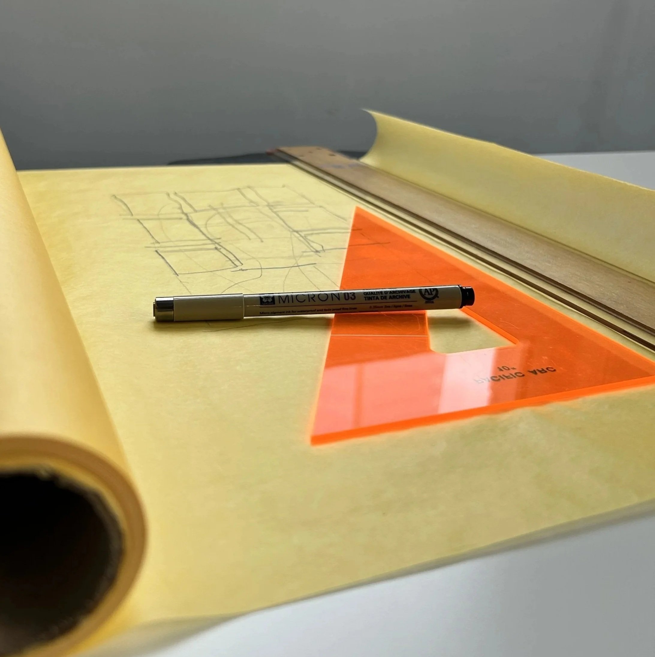 Open yellow sketchbook with architectural drawings, a black micron pen, orange transparent triangle set square on page, and metal ruler on the right.