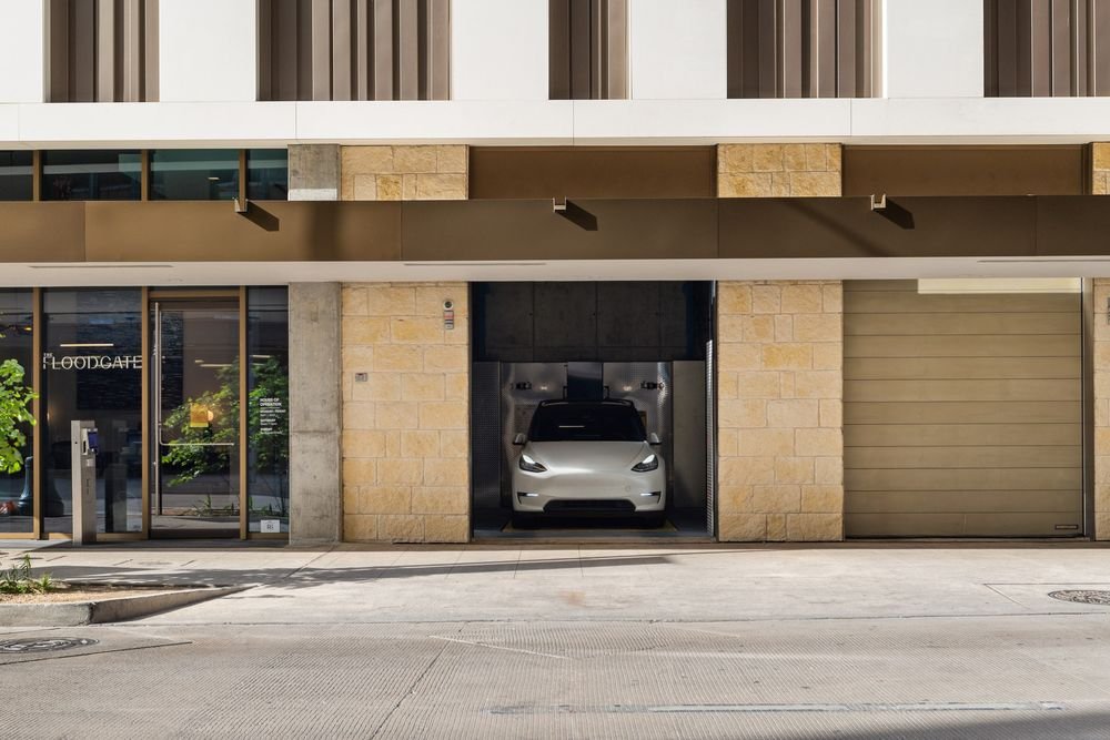 A silver Tesla parked in a glass-enclosed garage with a beige brick exterior, part of a modern commercial building, with a closed roll-up door to the right and the building's entrance on the left.