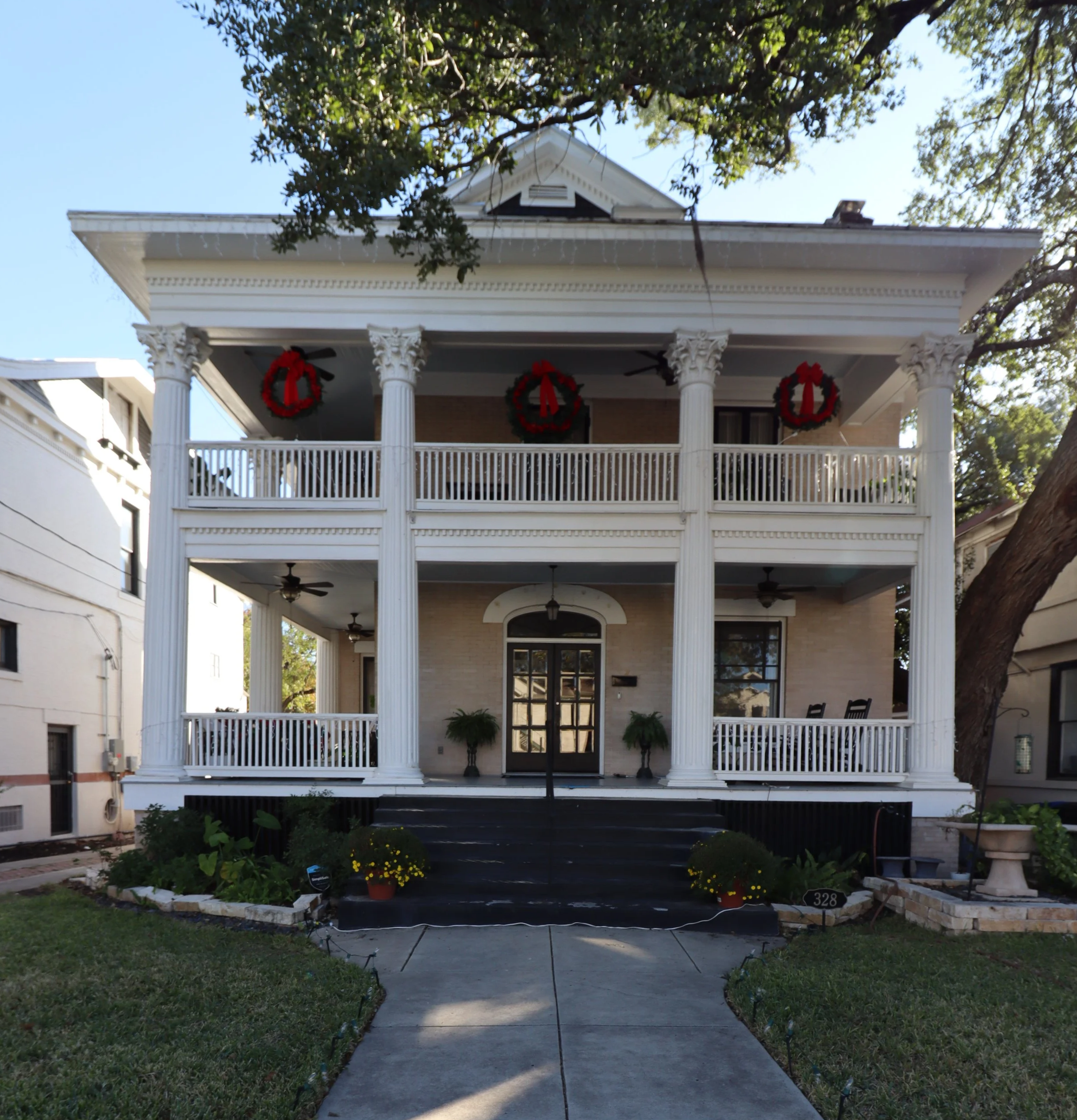 Front view of a large, two-story white house with decorated holiday wreaths on the upper balcony, black steps leading to the front door, surrounded by a well-maintained lawn and garden.