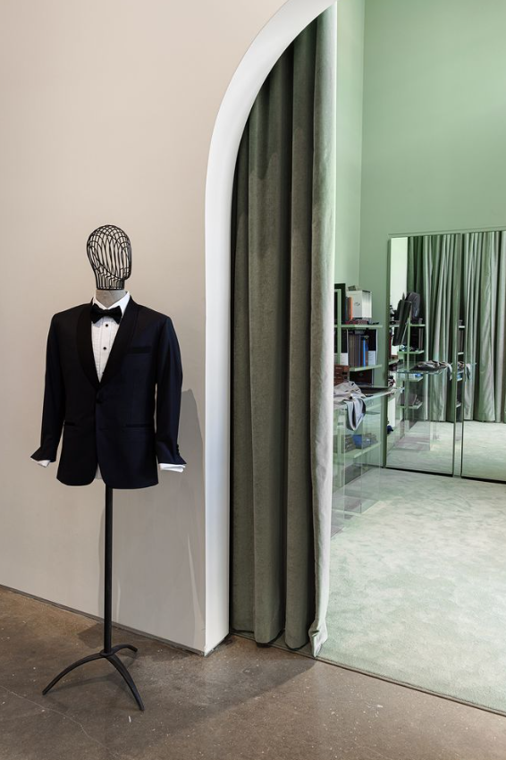 Mannequin dressed in a black tuxedo and bow tie, positioned near an arched doorway with green curtains in a room with a mirror and bookshelf.