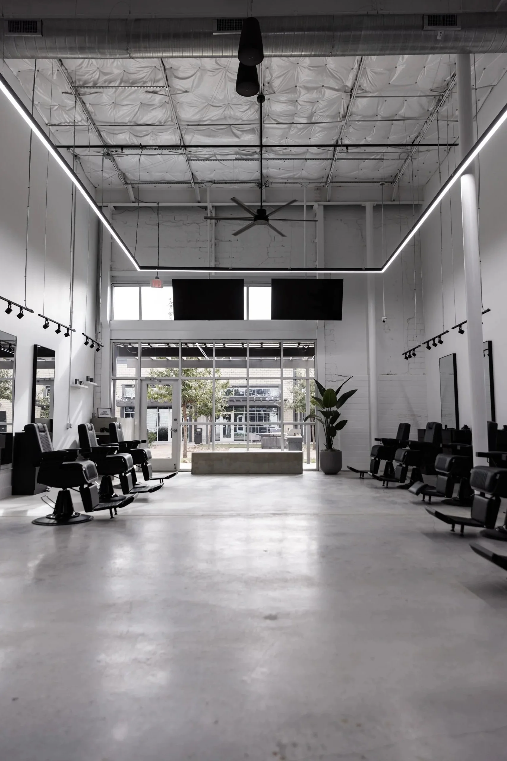 Modern, minimalistic salon interior with black chairs, large windows, hanging lights, ceiling fan, and a potted plant, illuminated by natural and LED lighting.