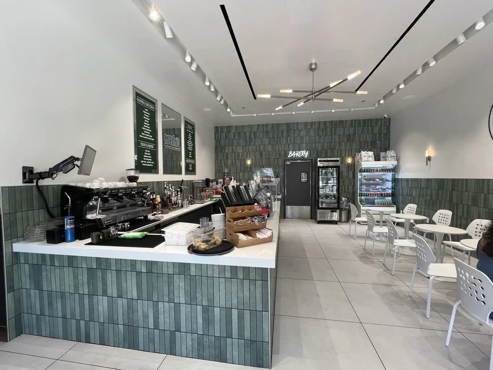 Modern coffee shop interior with a counter, coffee equipment, pastries, and seating area with white chairs and tables.