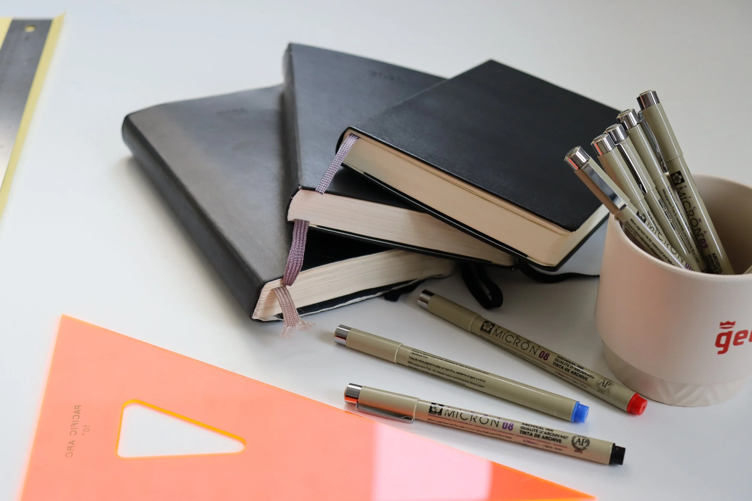 Desk with two black notebooks, a cup holding several gray Micron pens, orange plastic ruler, and a white surface background.