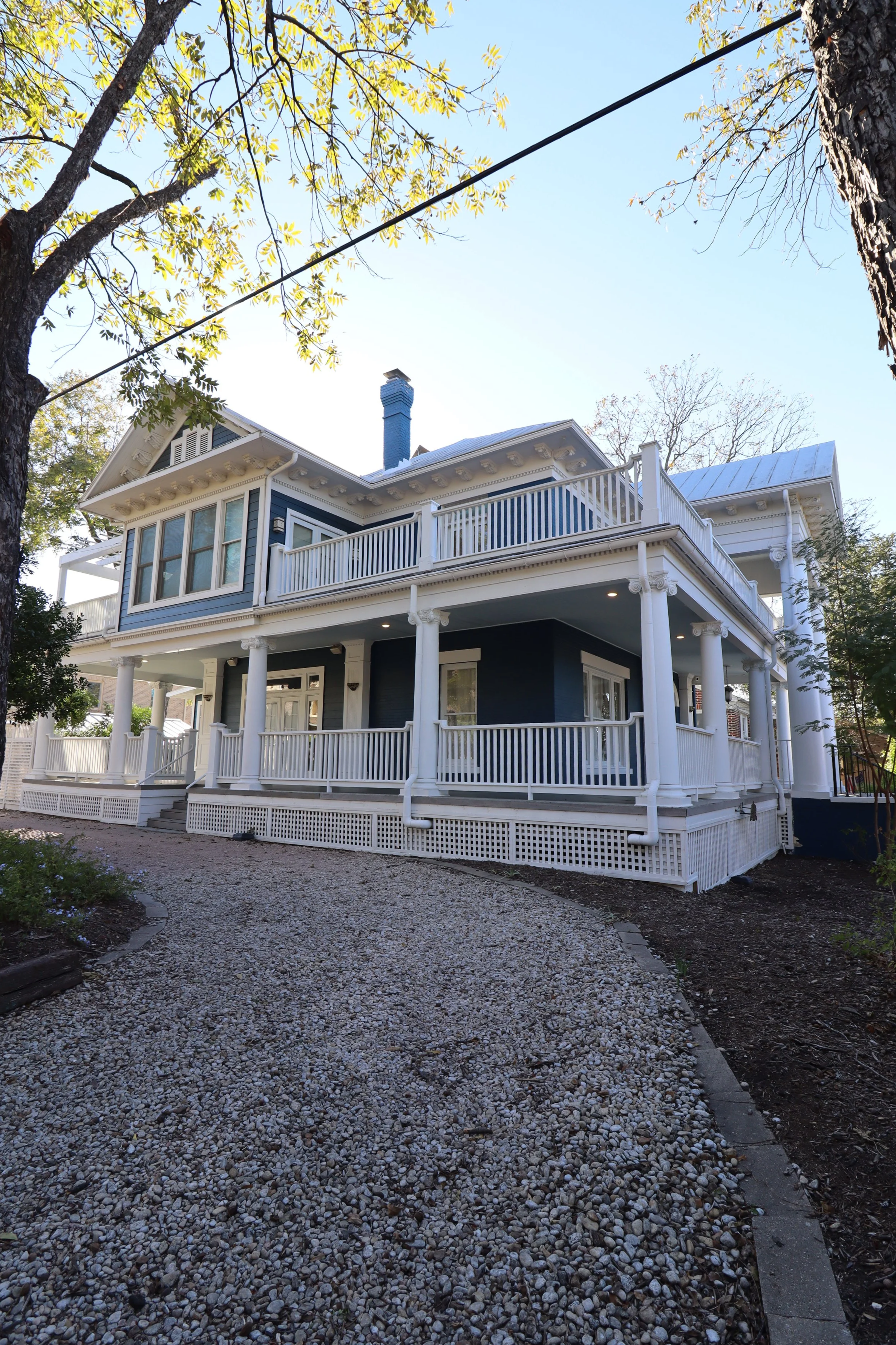 A large, two-story house with blue exterior and white trim, featuring multiple columns, balconies, and a gravel pathway leading to the entrance, surrounded by trees.