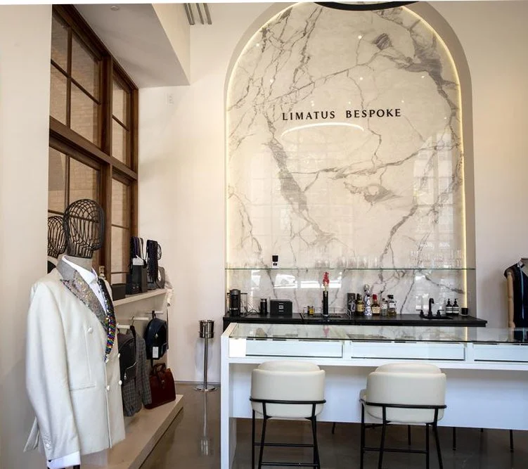 Interior of a modern boutique with a marble wall feature and a white bar counter, labeled 'LIMATUS BESPOKE', with two white chairs in front of it, and clothing mannequins displaying suits and accessories.