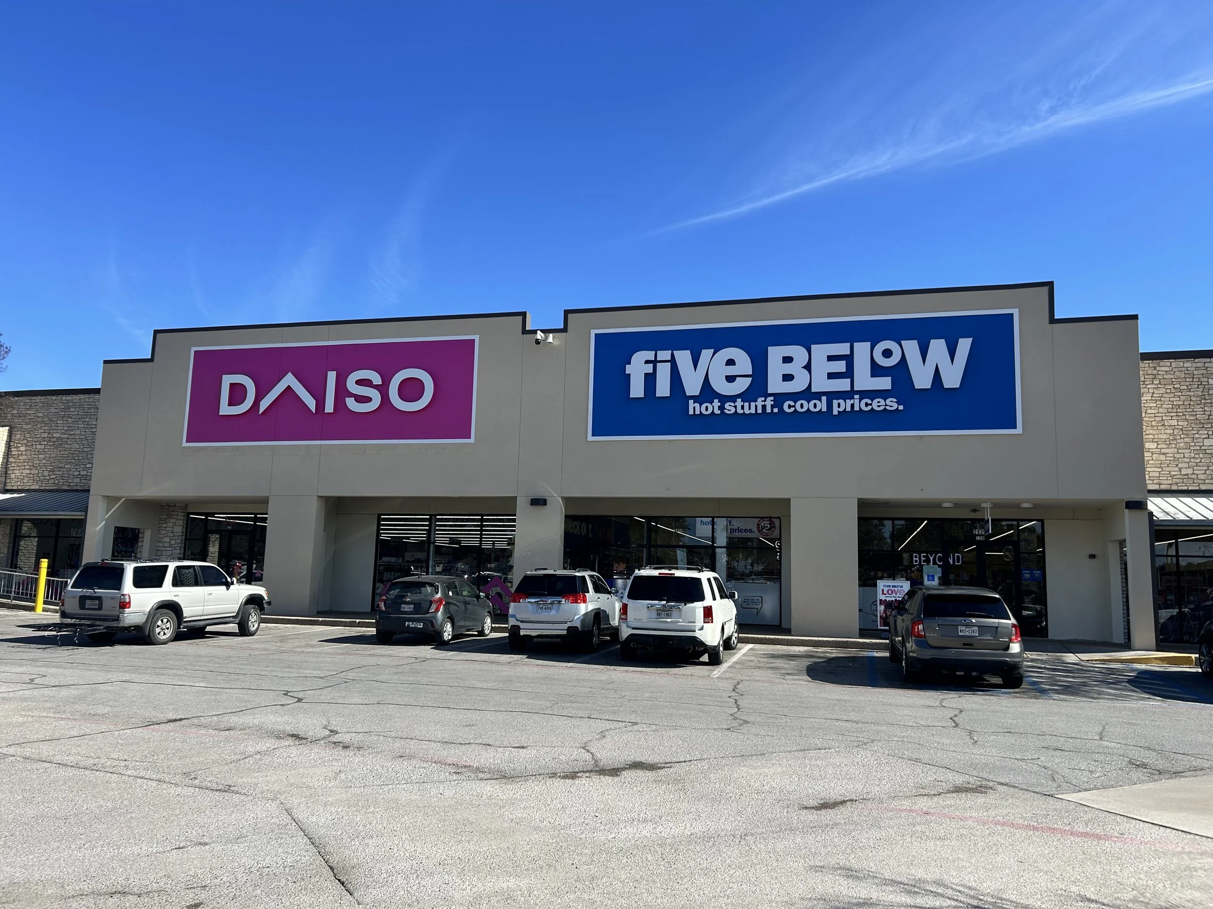 Exterior of a shopping plaza with two large signs, one for Daiso and one for five BELOW, with parked cars in front on a clear day with blue sky and some clouds.
