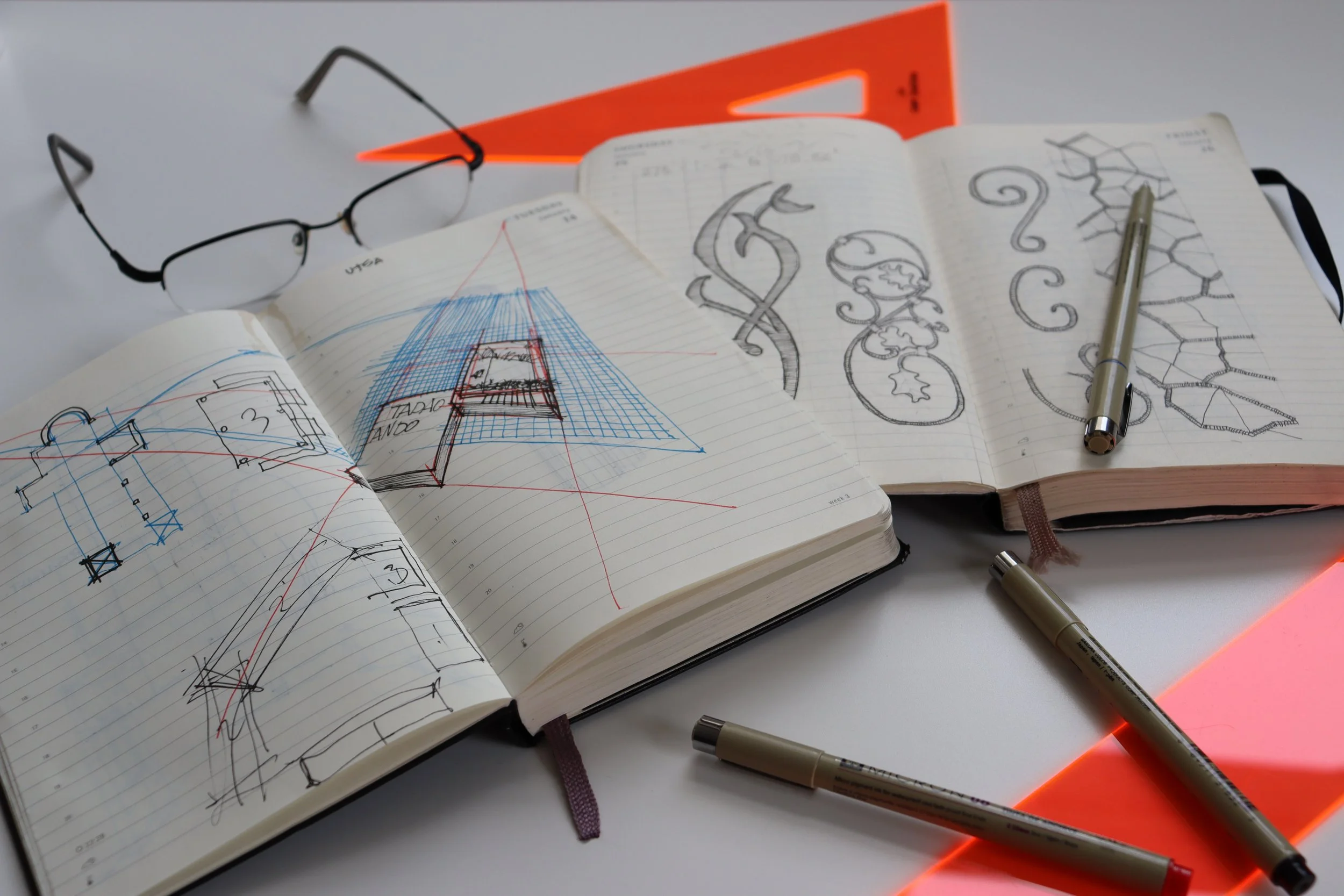 Open sketchbooks with architectural and decorative drawings, a pair of glasses, red triangular set square, and three metallic pens on a white desk with glowing orange edges.