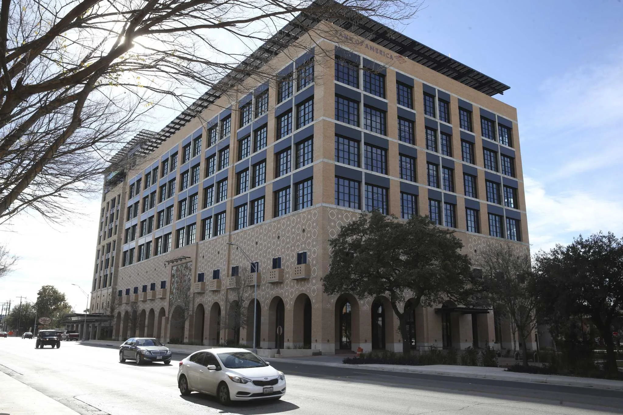 A multi-story modern building with large windows and decorative brickwork, situated along a city street with cars parked and trees lining the sidewalk.