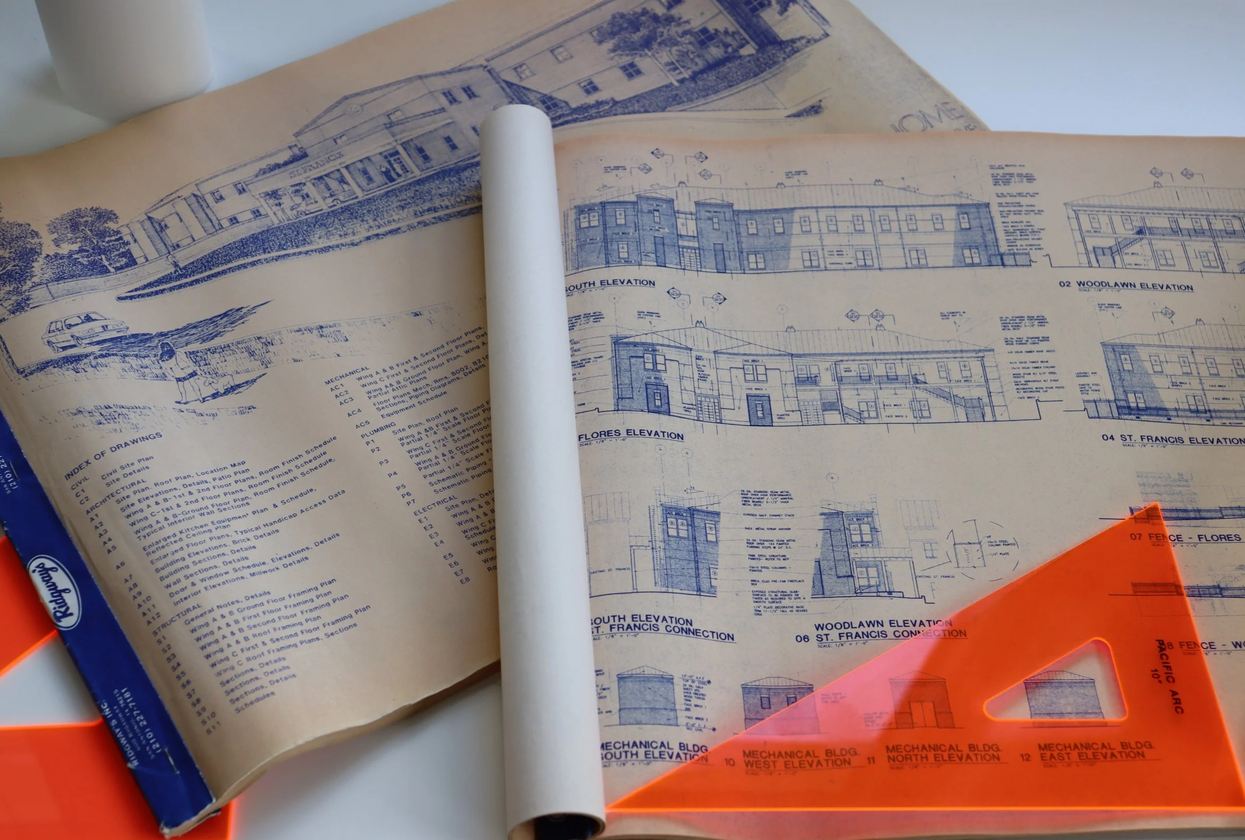 Blueprints of a building with architectural plans, elevation views, and notes, partially covered by a rolled-up section and a translucent orange triangle ruler.