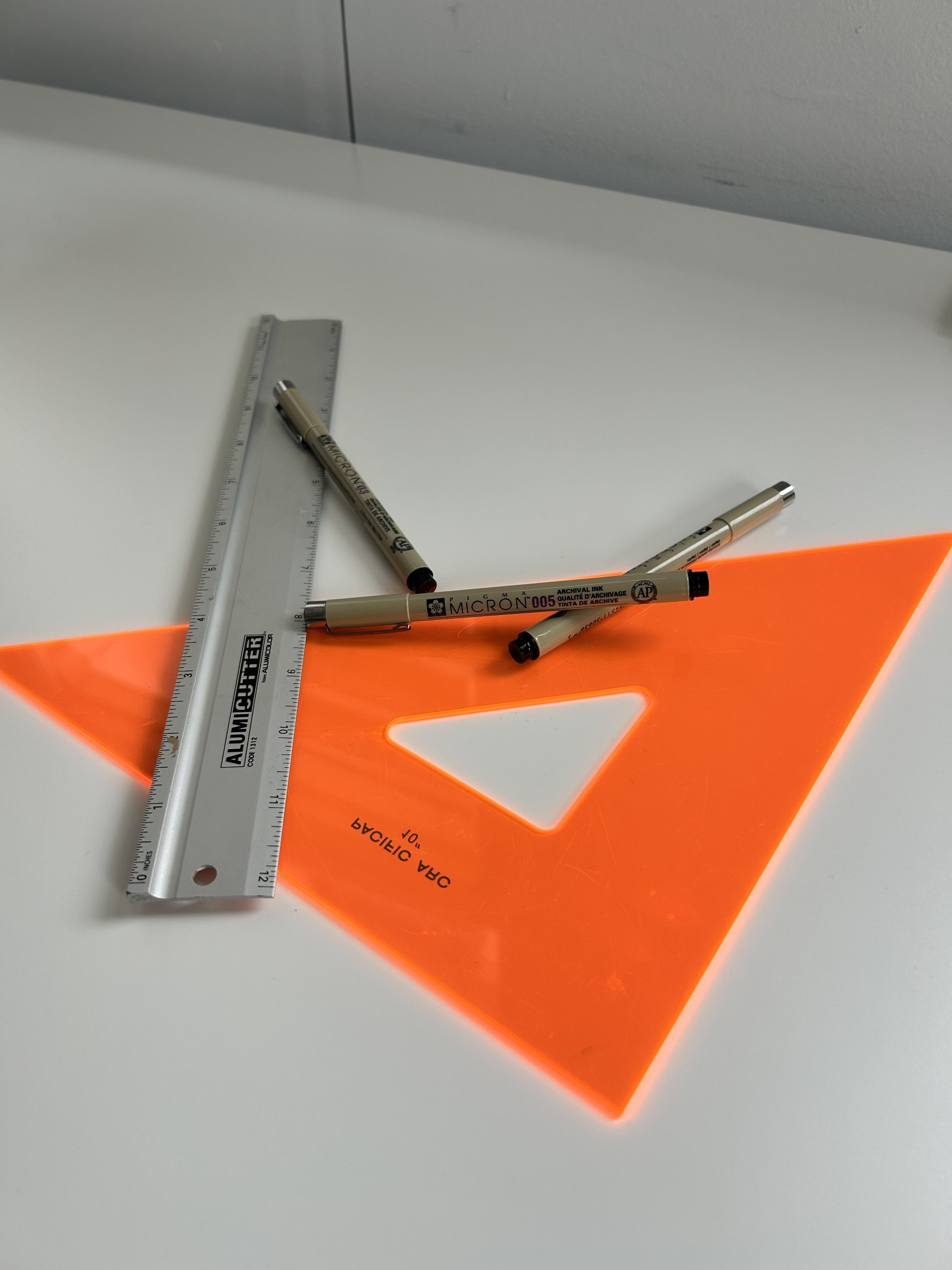 An orange translucent geometric drafting triangle with a triangular cutout, a metal ruler, and three black and gold fine-tipped pens on a white surface.
