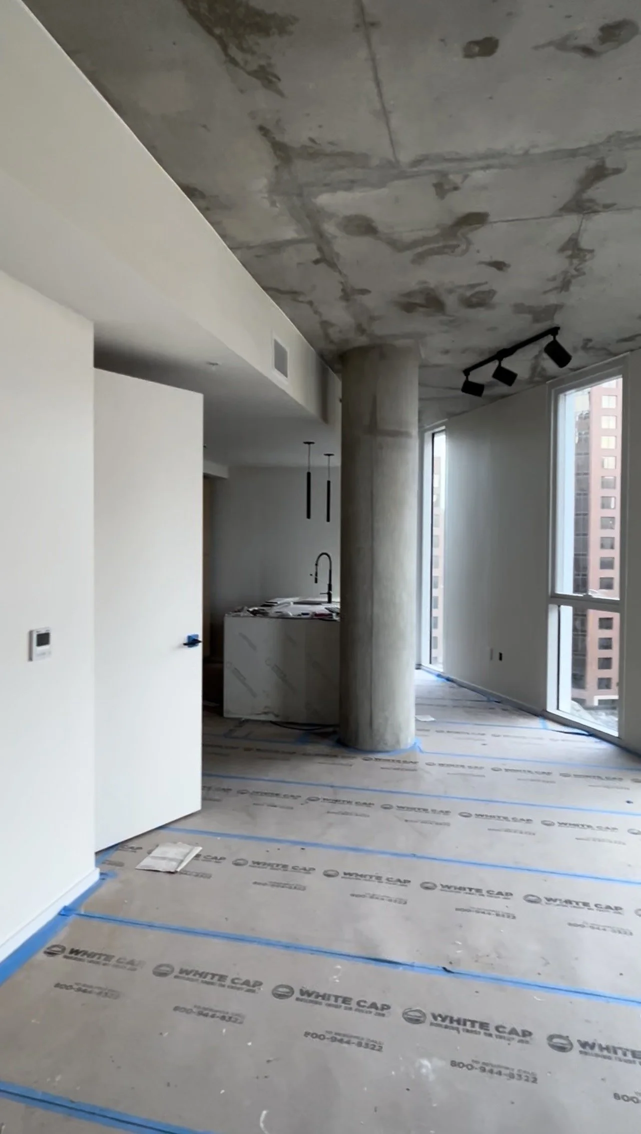 Interior of an unfinished high-rise apartment with concrete ceiling and floor, large windows, partially painted walls, and a concrete column in the living area.