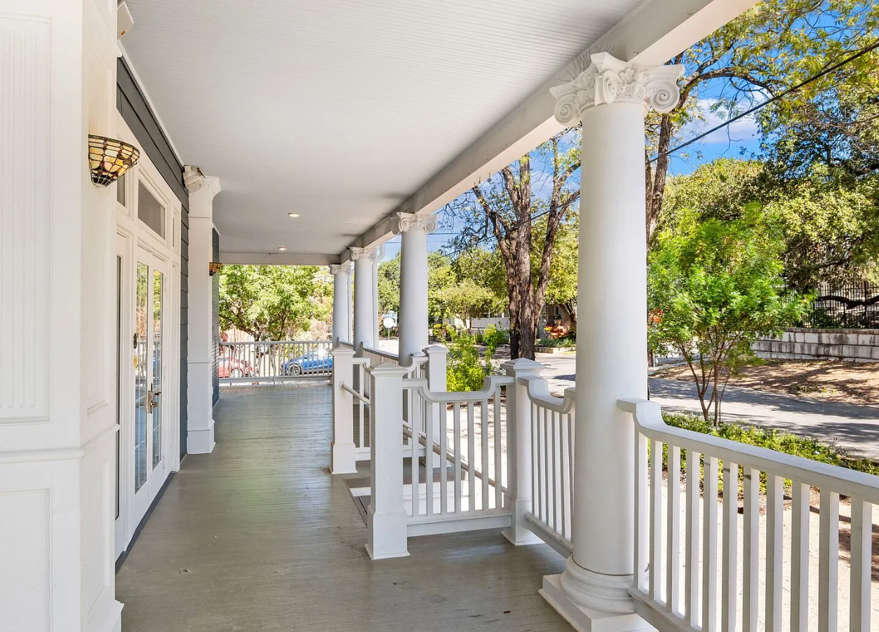 Empty front porch with white columns, railing, and door, overlooking a street with trees and parked cars.