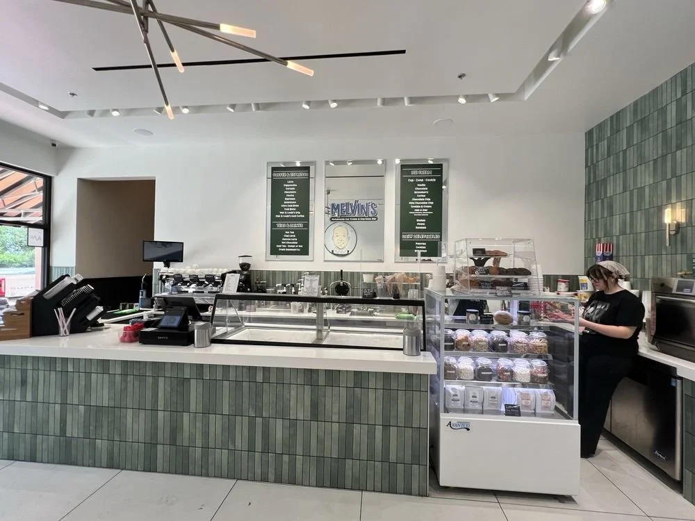 Interior of a modern ice cream shop with a green tiled front counter, digital menu boards, a refrigerated display case filled with pre-packaged ice cream, and a staff member wearing a hairnet working behind the counter.