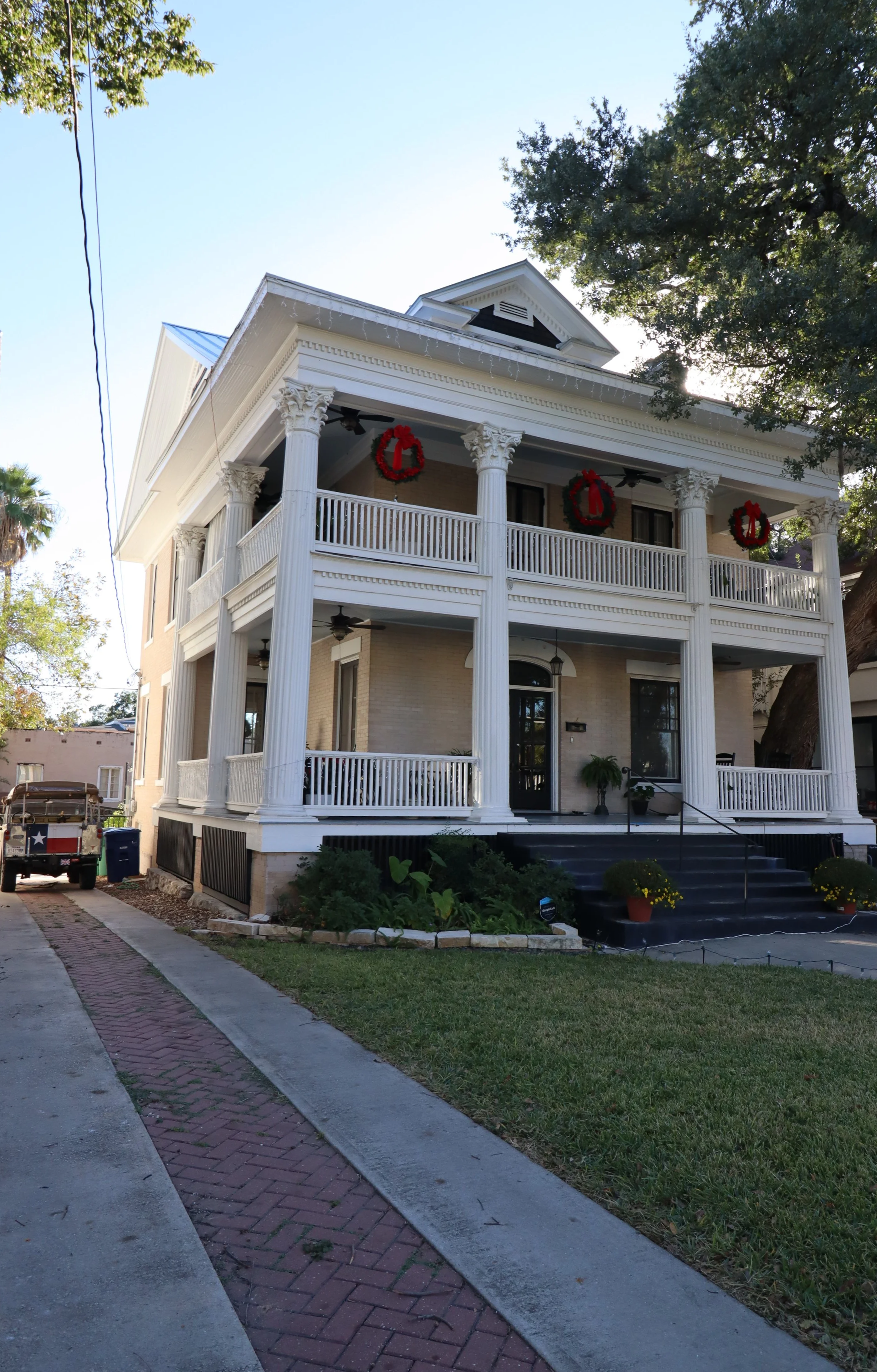 A large, three-story house with a front porch supported by white columns, decorated with Christmas wreaths. The house has a staircase leading to the porch, potted plants, and a small garden in front. A tree is on the right side, and a driveway with a truck is on the left.