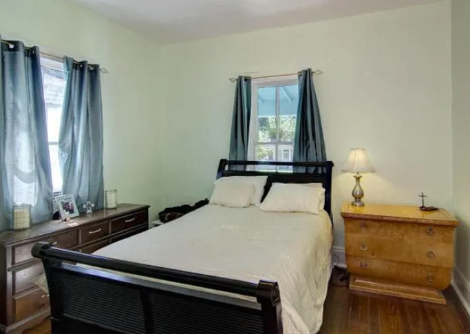 A bedroom with a bed, dark wood headboard and footboard, white bedding, two pillows, two blue curtains on windows, a wooden dresser with framed photos and candles, a nightstand with a lamp, and hardwood flooring.