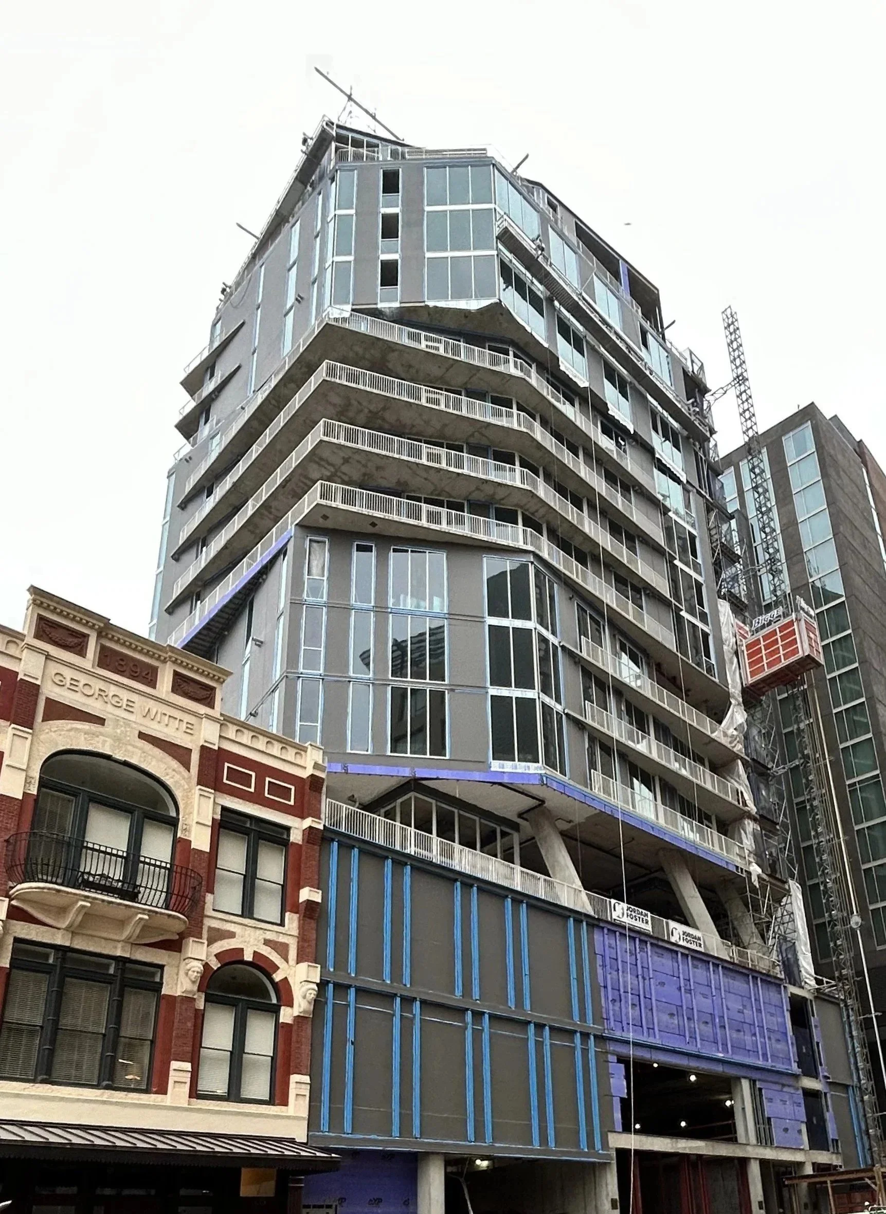A multi-story modern building under construction with glass windows, balconies, and construction equipment, next to an older historic building with decorative brickwork.