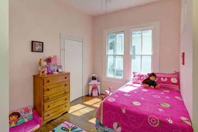A child's bedroom with pink walls, a wooden dresser, a bed with pink floral bedding, and a plush monkey toy. The room has a window letting in natural light and a small bookshelf or toy organizer near the window.