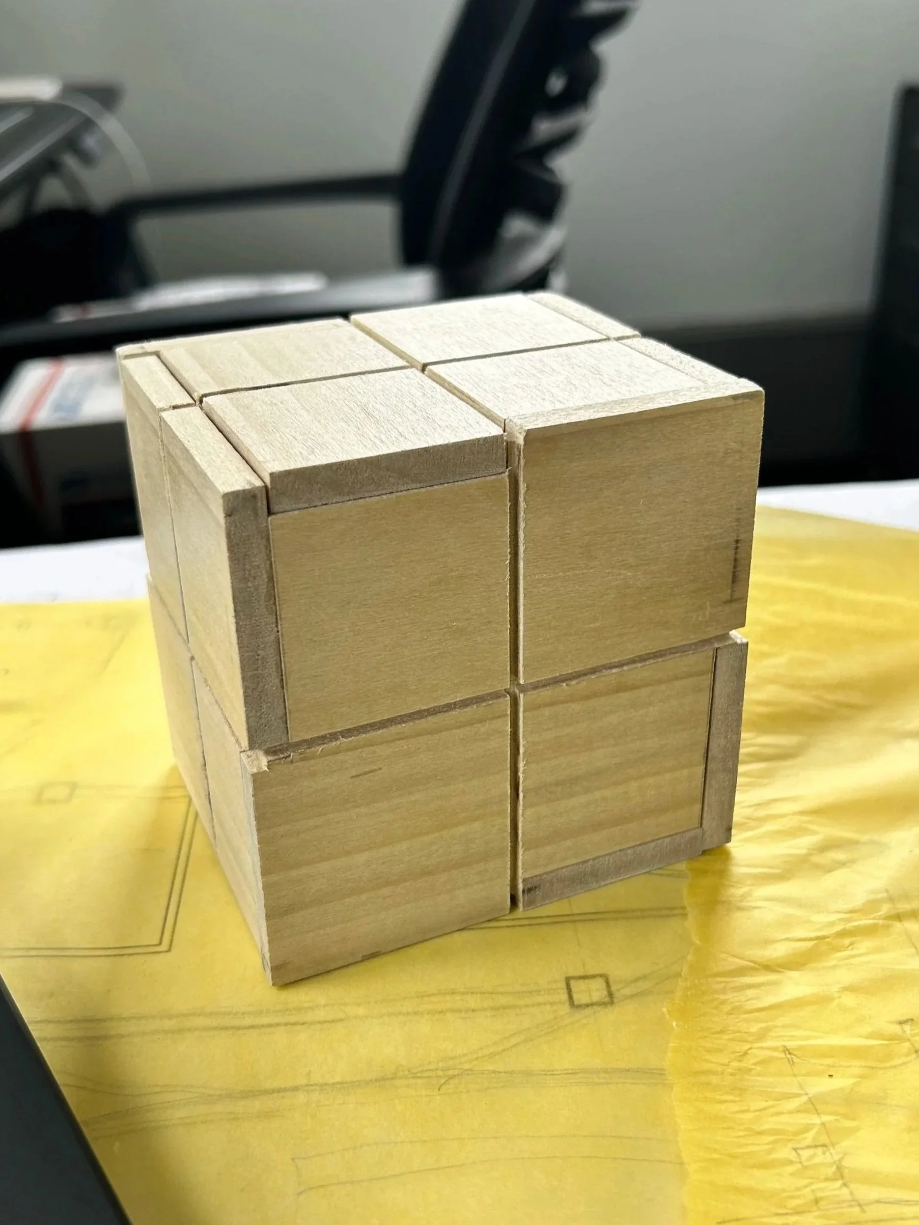 A wooden puzzle cube with interlocking pieces, resting on a yellow surface with a diagram or pattern.