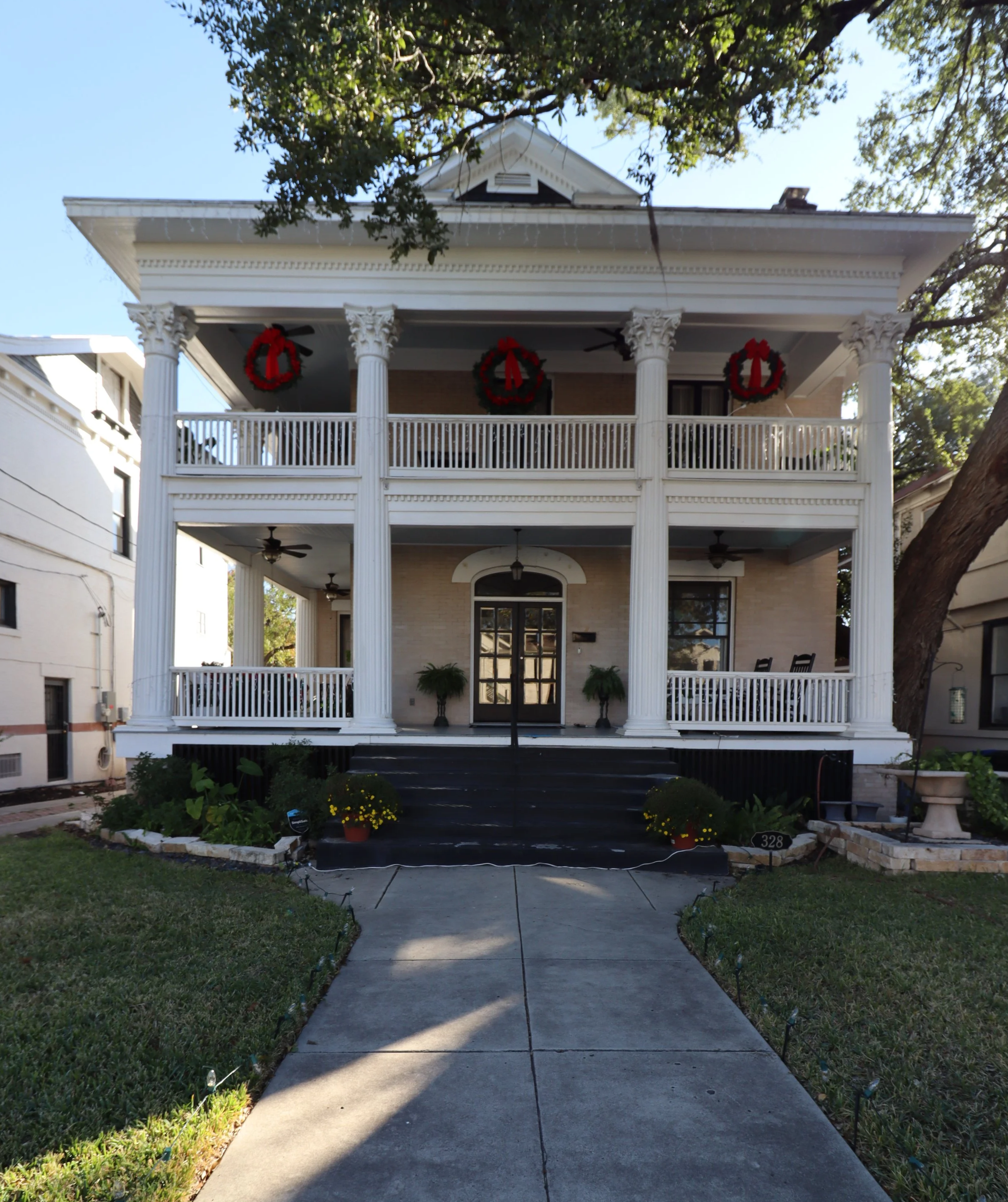 A large, two-story house decorated for Christmas with red wreaths and greenery on the front porch. The house has white columns, a black front door, and a driveway leading up to it.