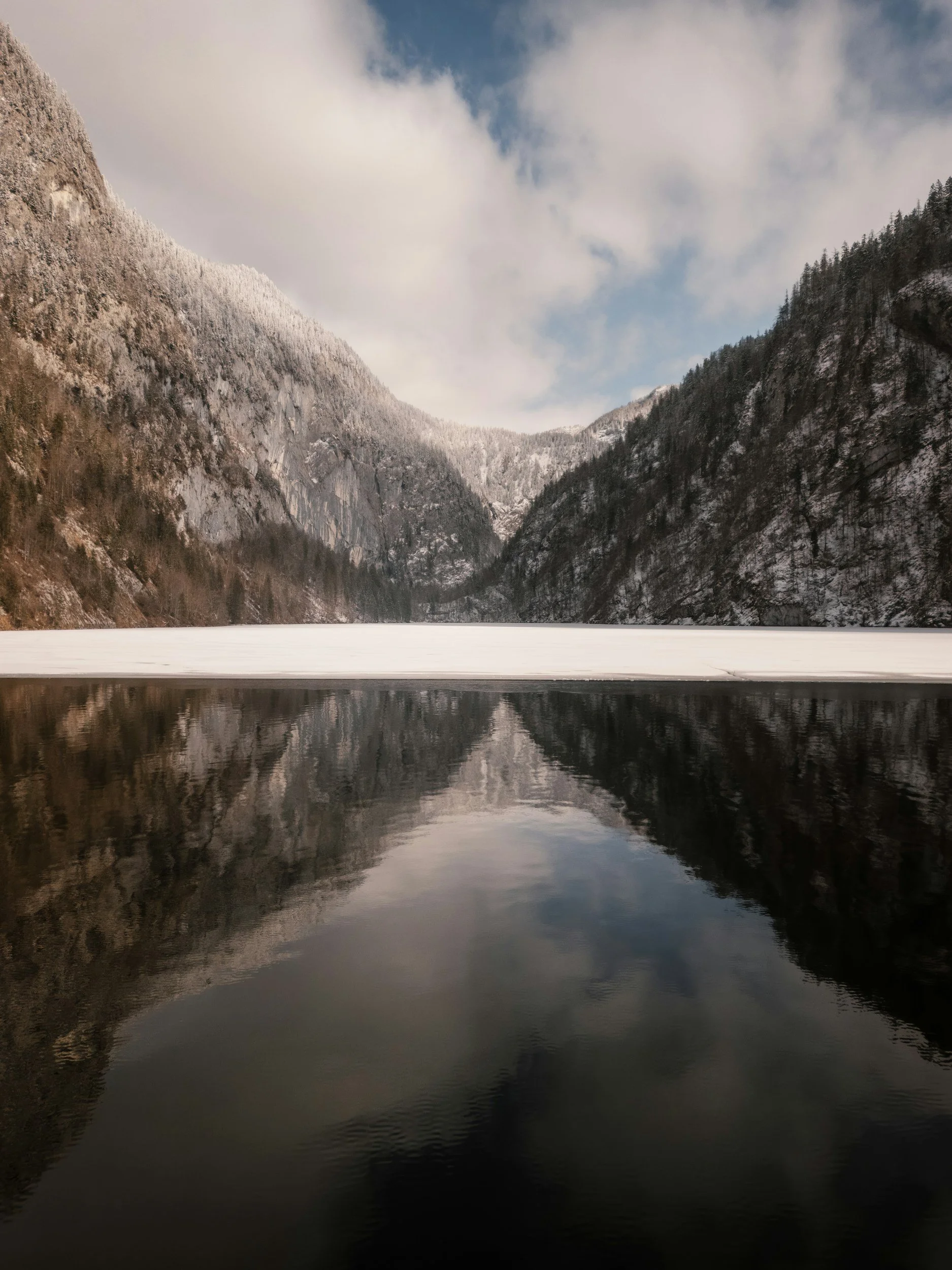 A winter landscape showing snow-capped mountains on either side of a partially frozen lake with a reflection of the mountains and sky in the water.
